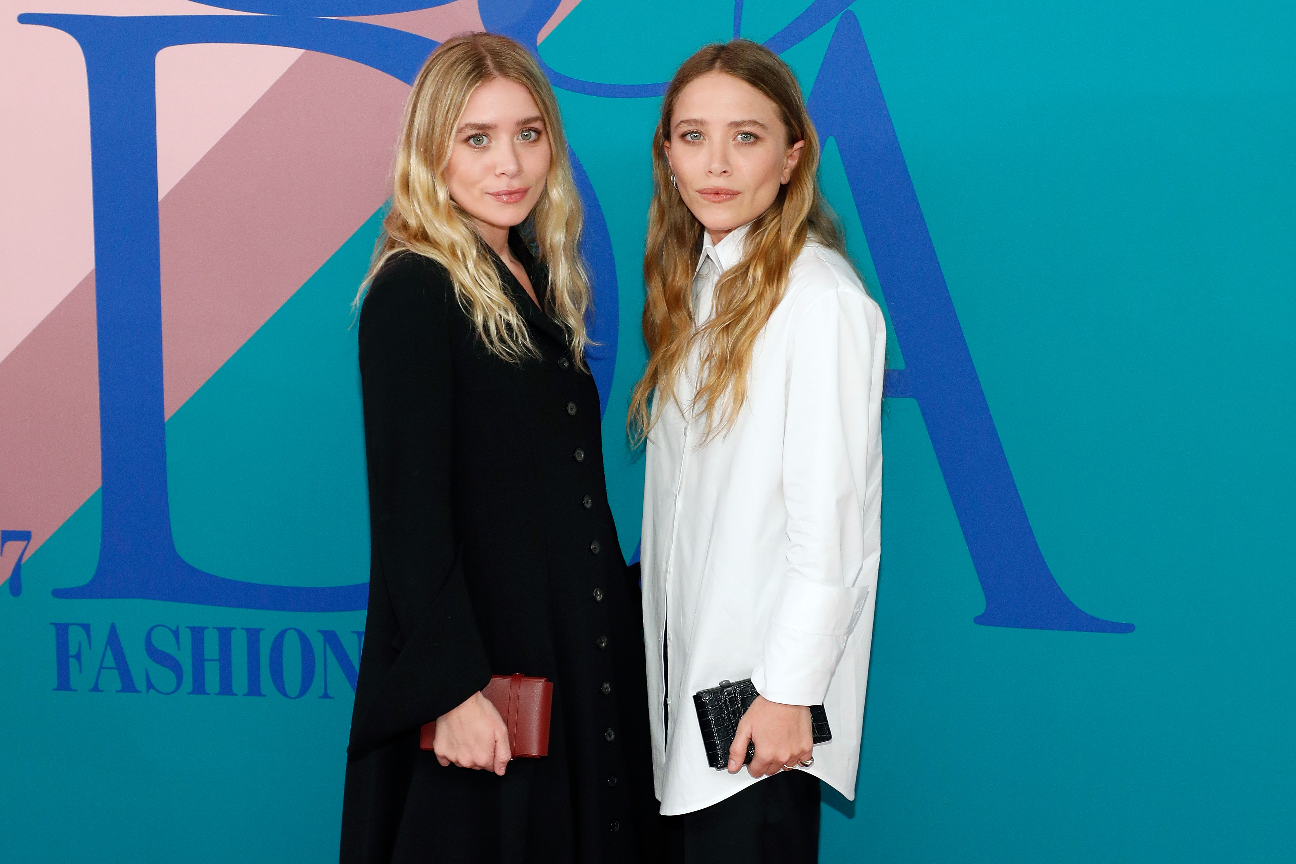 Mary-Kate and Ashley Olsen stand together at a fashion event, facing forward with neutral expressions. One wears a dark coat, the other a light blouse, both holding small clutches.