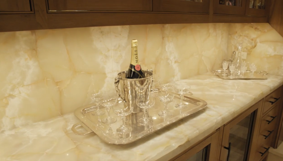A close-up view highlights the illuminated marble backsplash and countertop of the theater bar, where a bottle of champagne rests in a polished silver ice bucket. Silver serving trays display delicate crystal glassware and decanters beneath built-in wooden cabinetry. | Source: YouTube/Architectural Digest