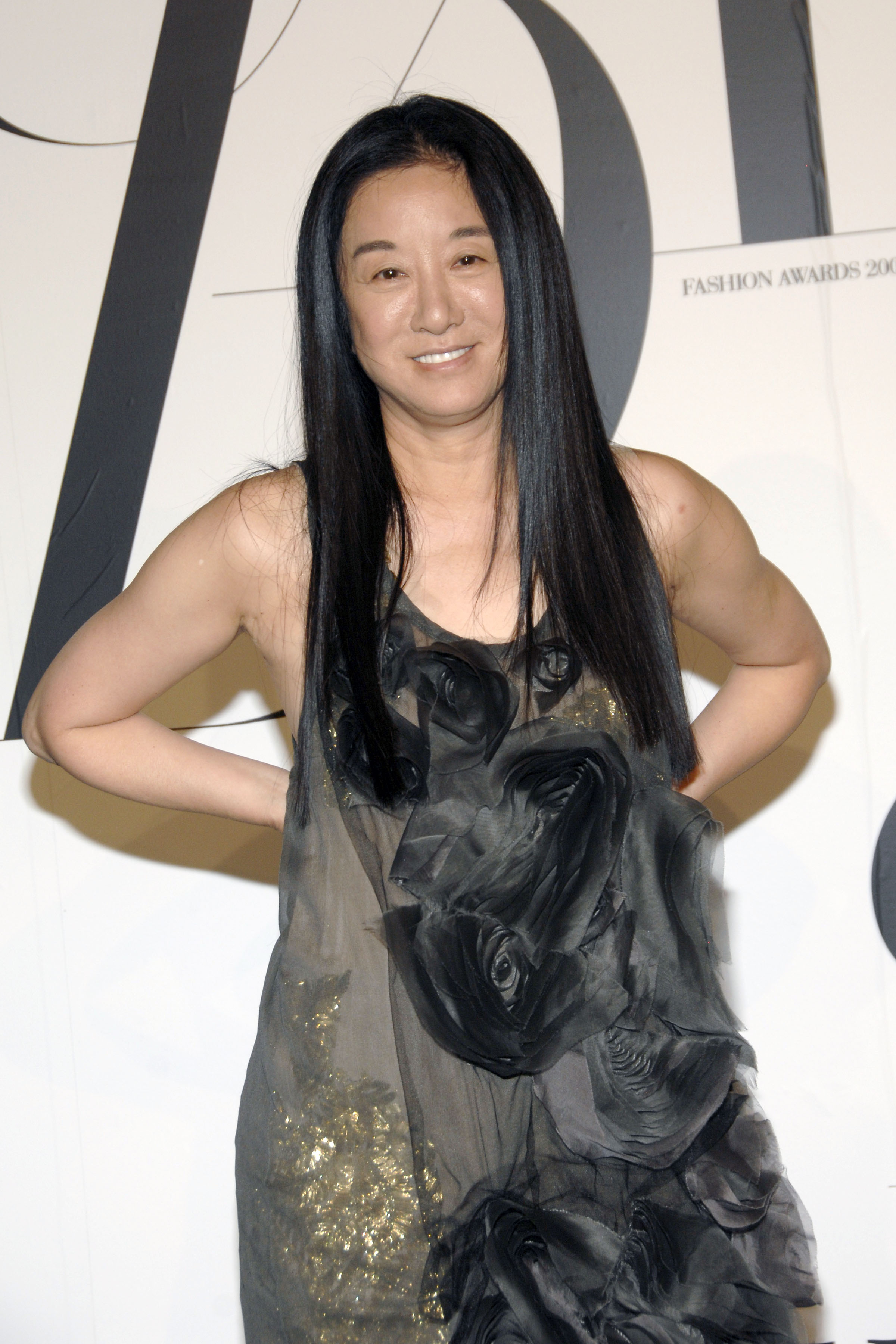 Vera Wang arrives at the CFDA Awards wearing a sheer black gown adorned with large floral appliqués and a dusting of gold shimmer. With her hands on her hips and a subtle smile, she exudes quiet confidence in front of the press wall.