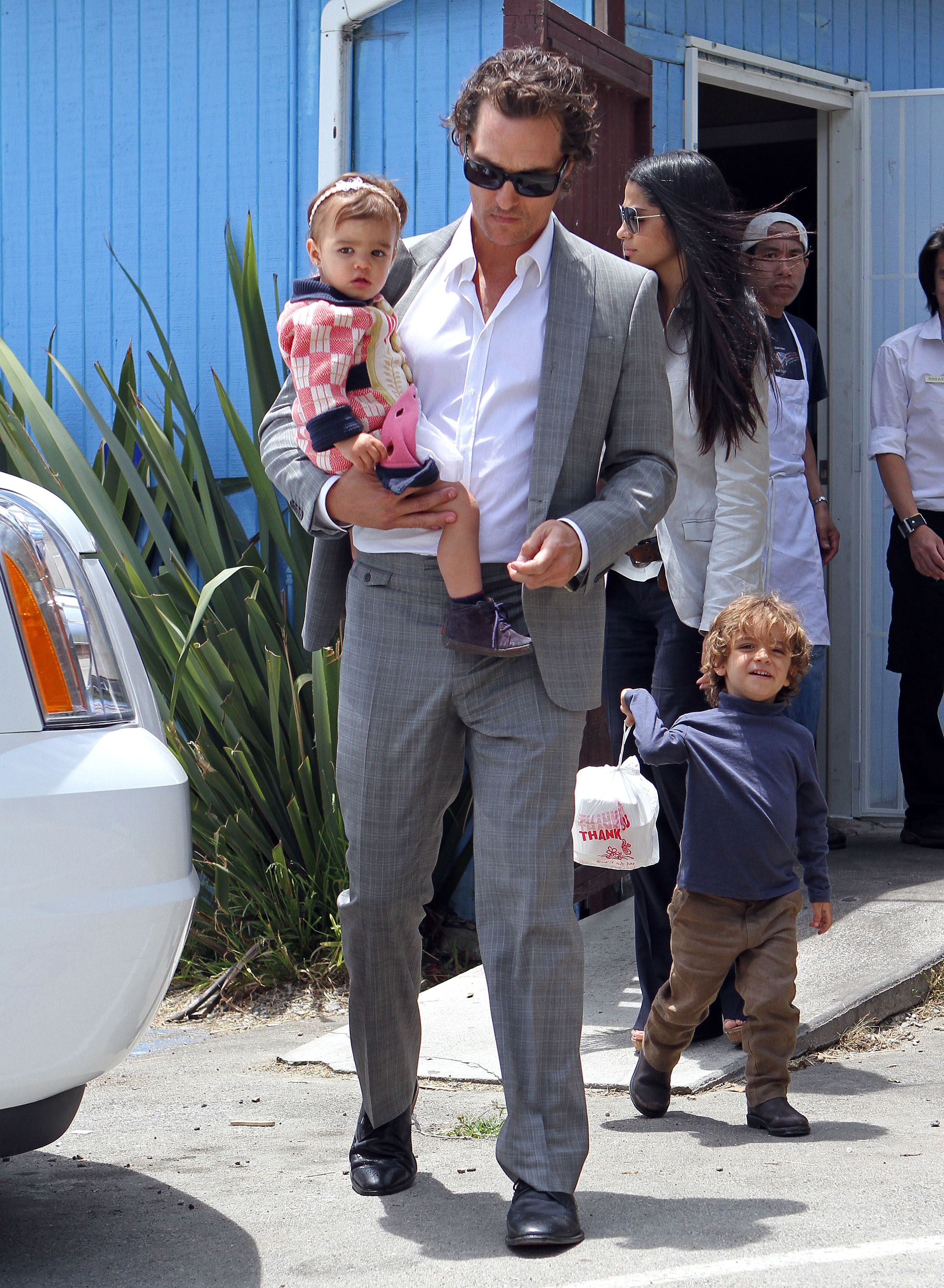 Vida Alves McConaughey is carried in Matthew McConaughey’s arms. Levi Alves McConaughey walks beside them holding a small bag. Camila Alves follows behind as they move along a sidewalk.