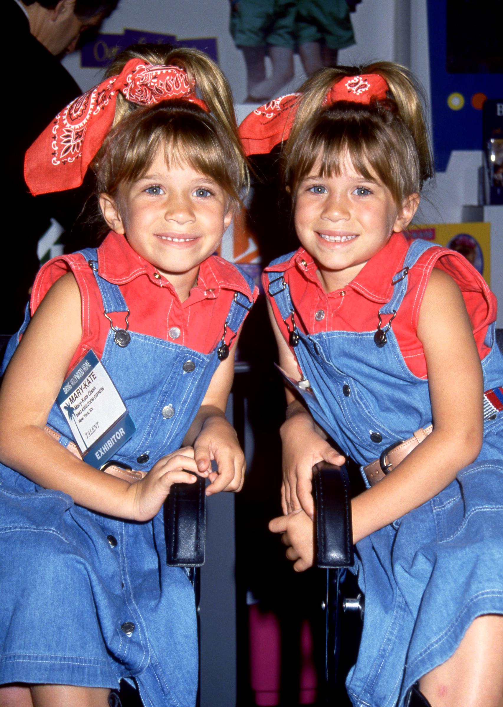 Mary-Kate and Ashley Olsen lean in close, smiling brightly in red tops and denim overalls. Matching bandanas and their pose give the moment a playful feel.