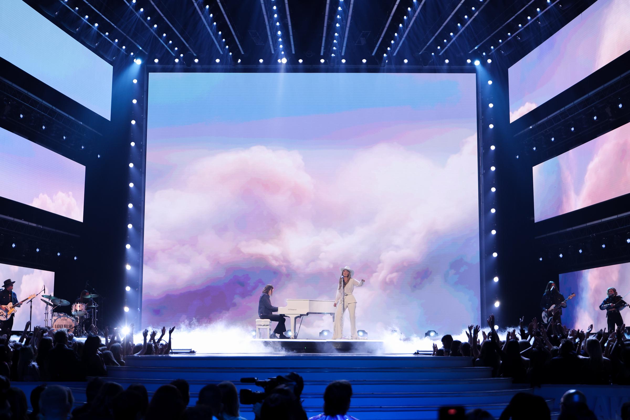 Wilson stands on stage alongside a pianist, the backdrop behind her a cloudy, ethereal swirl that gives the performance a dreamlike feel. With guitarists and other band members positioned on either side, the scene is both intimate and atmospheric.
