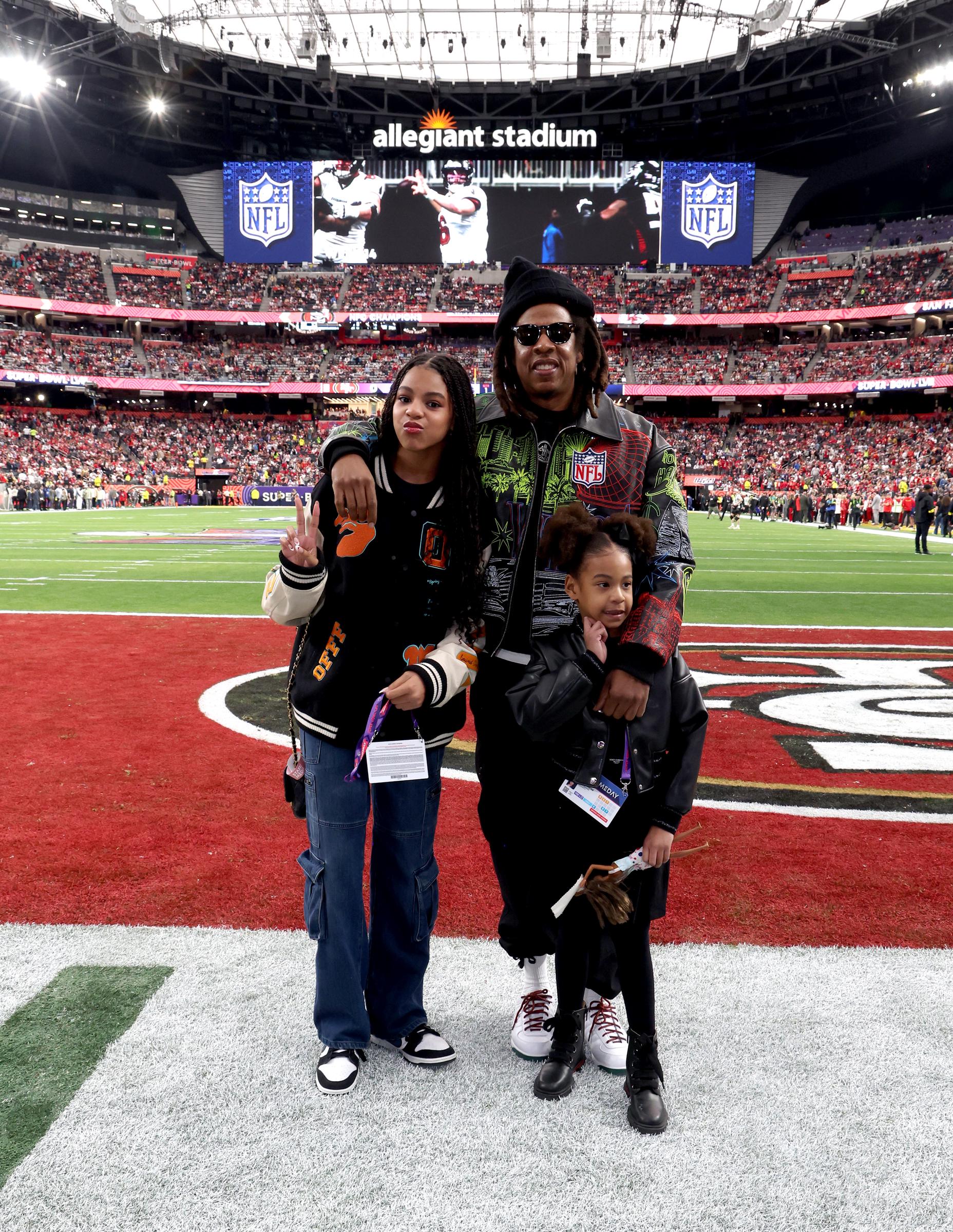 Jay-Z stands on the field with Blue Ivy and Rumi, his arm around both daughters as they pose near the end zone. The trio appears relaxed against the packed stadium backdrop.