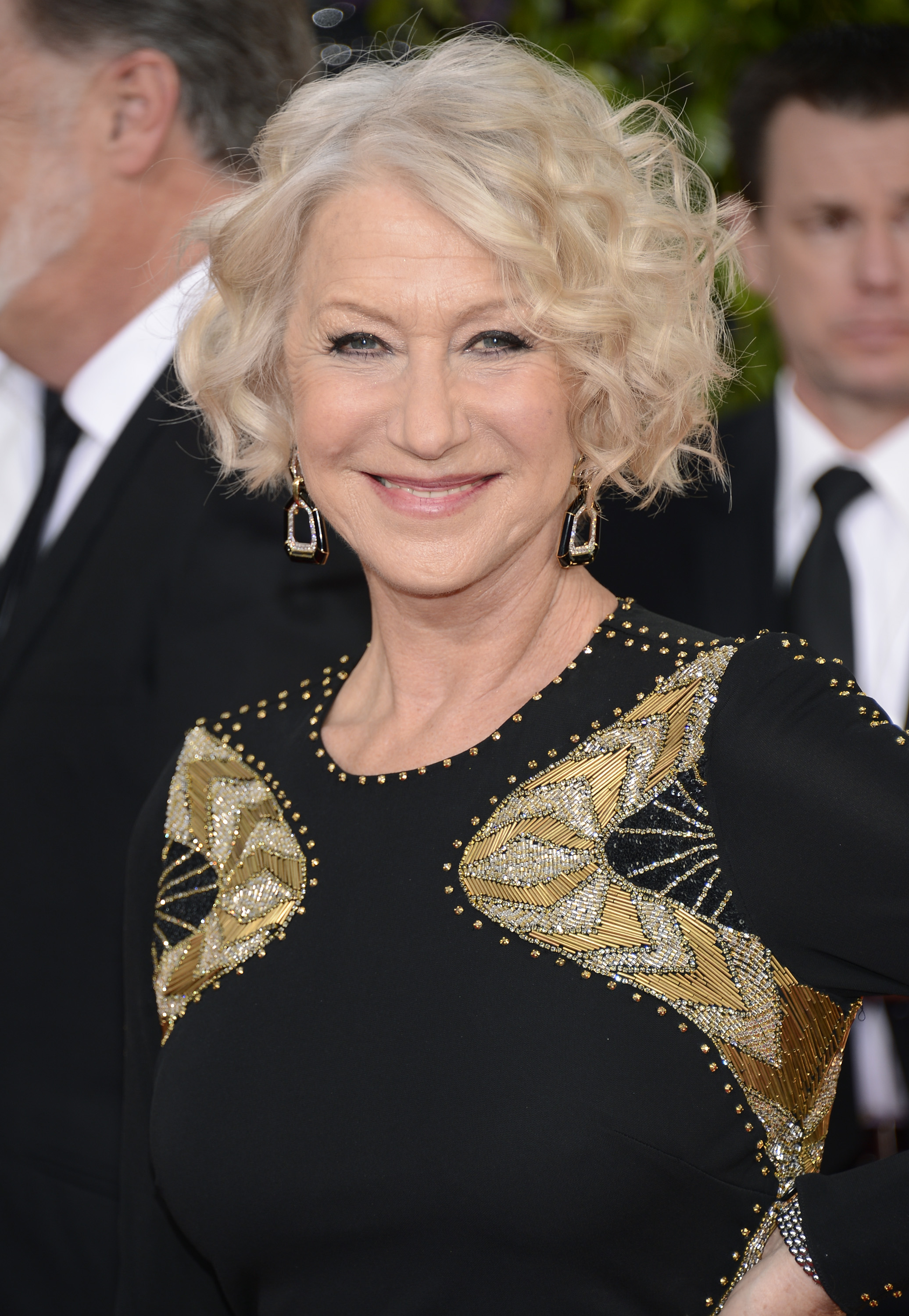 Helen Mirren radiates confidence on the red carpet in a black gown adorned with intricate gold and silver beadwork across the shoulders, pairing the striking ensemble with geometric drop earrings and her signature platinum curls styled in soft, voluminous waves.