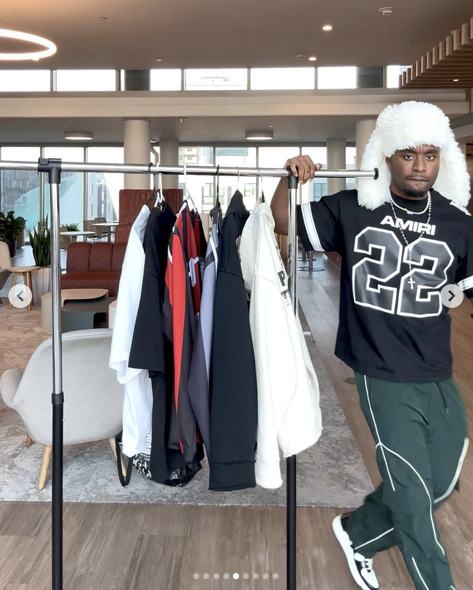 Philip Enewally is pictured standing next to a collection of hoodies, jerseys, and t-shirts from his clothing brand, shared on February 20, 2025 | Source: Instagram/p2isthename