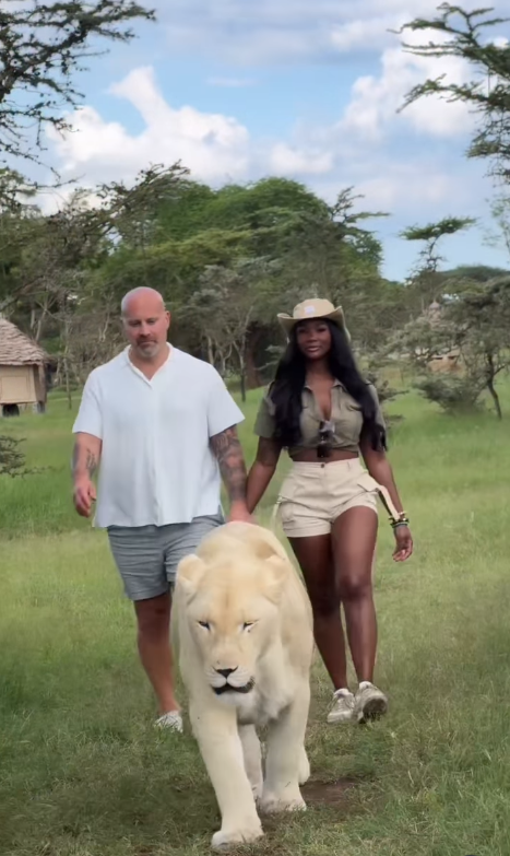 Ashly Robinson and Joe McCann walk side by side through open grassland, calmly accompanying a majestic white lion as the serene, safari-like landscape stretches out behind them. | Source: Instgaram/ashleejenae