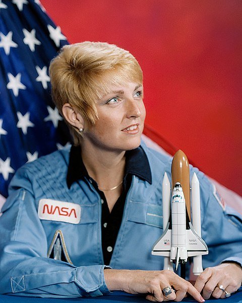 Millie Hughes-Fulford wearing NASA uniform with U.S. flag in background | Source: Wikimedia Commons
