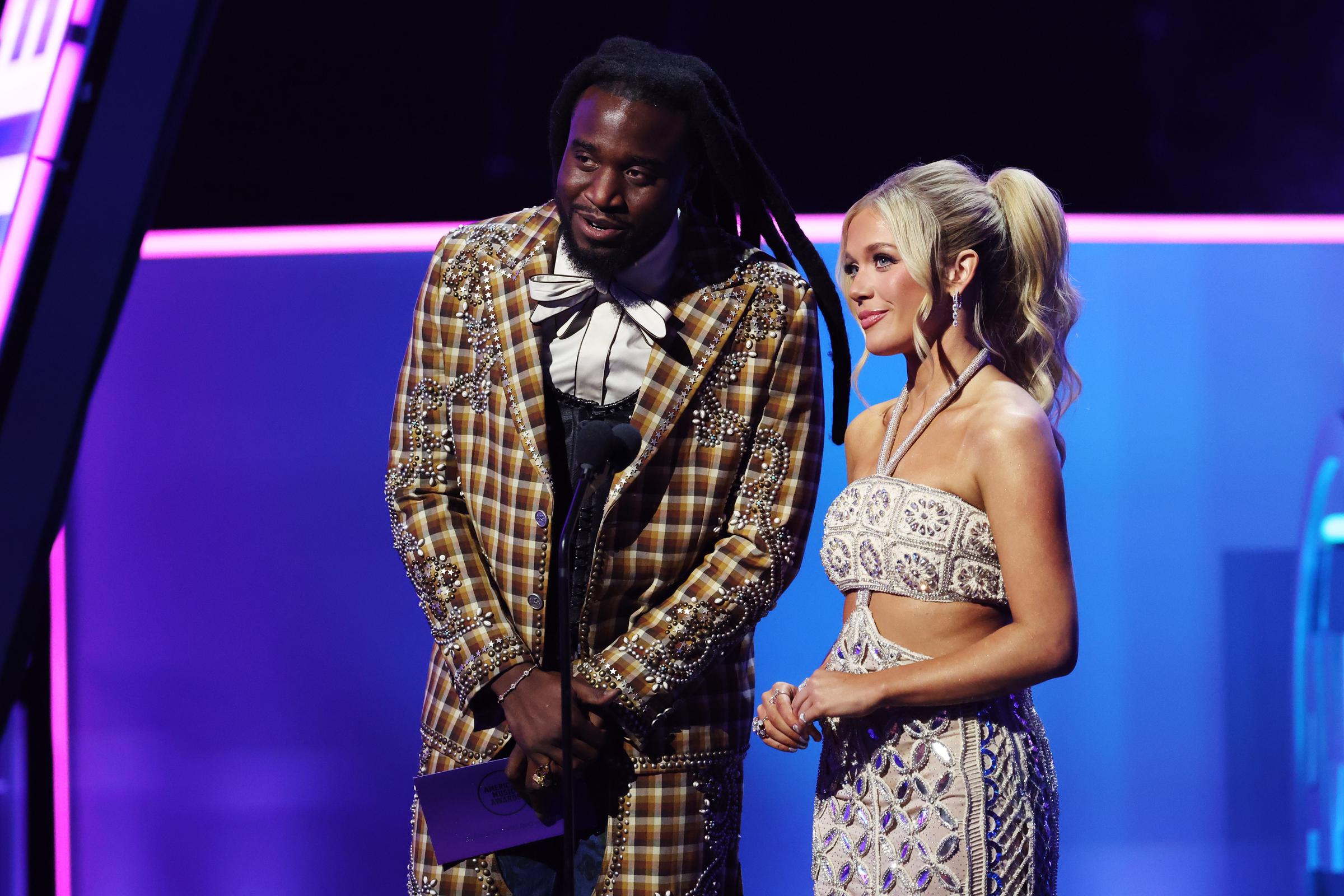 Side by side at the podium, Moroney listens intently with a poised smile as the night's next award is announced, her elegance complementing the flamboyant flair of her co-host.