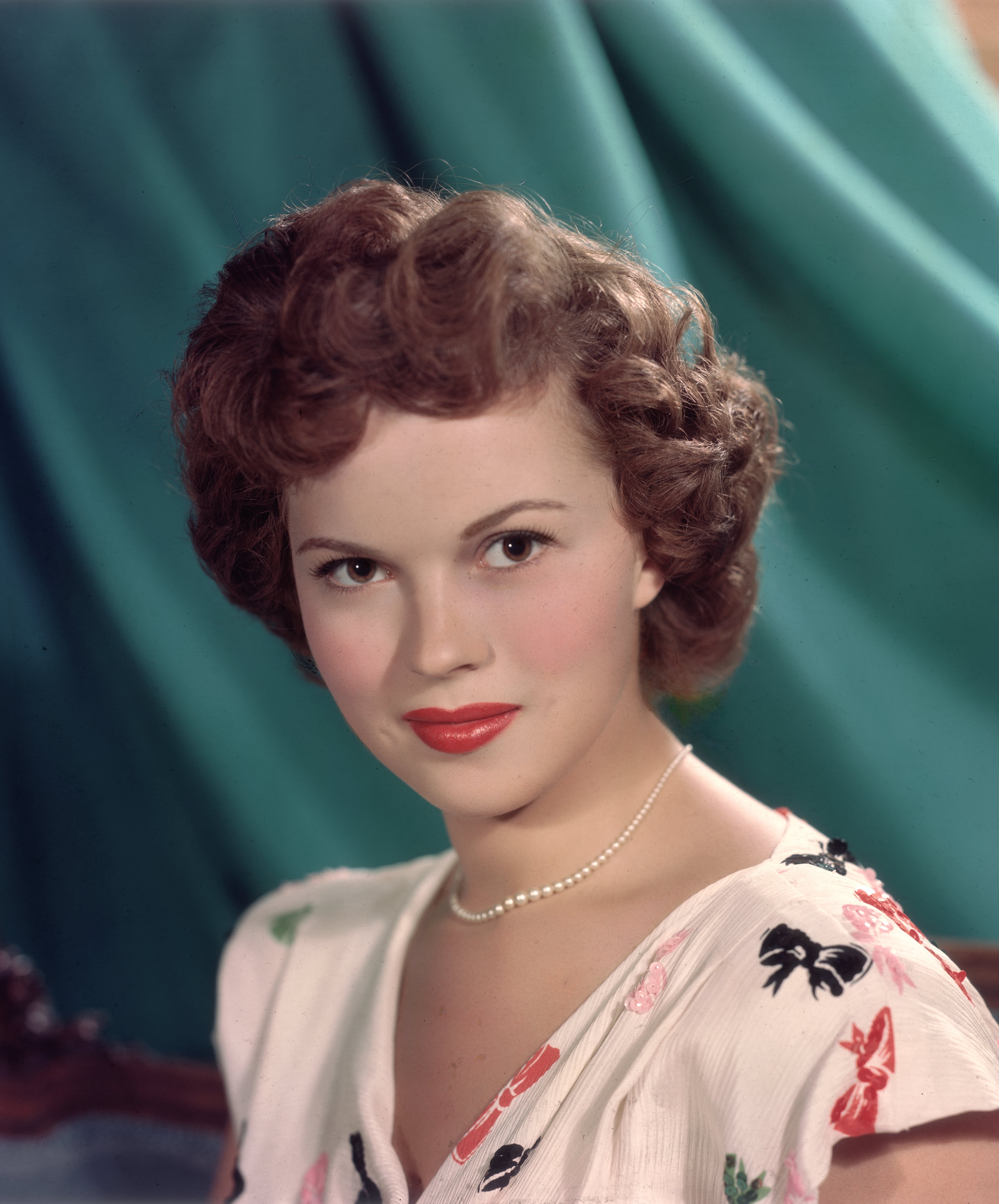  Headshot studio portrait of American actor Shirley Temple, in front of a green backdrop, wearing a short sleeve white blouse with a colored bows pattern and a pearl necklace. circa 1955 | Source: Getty Images