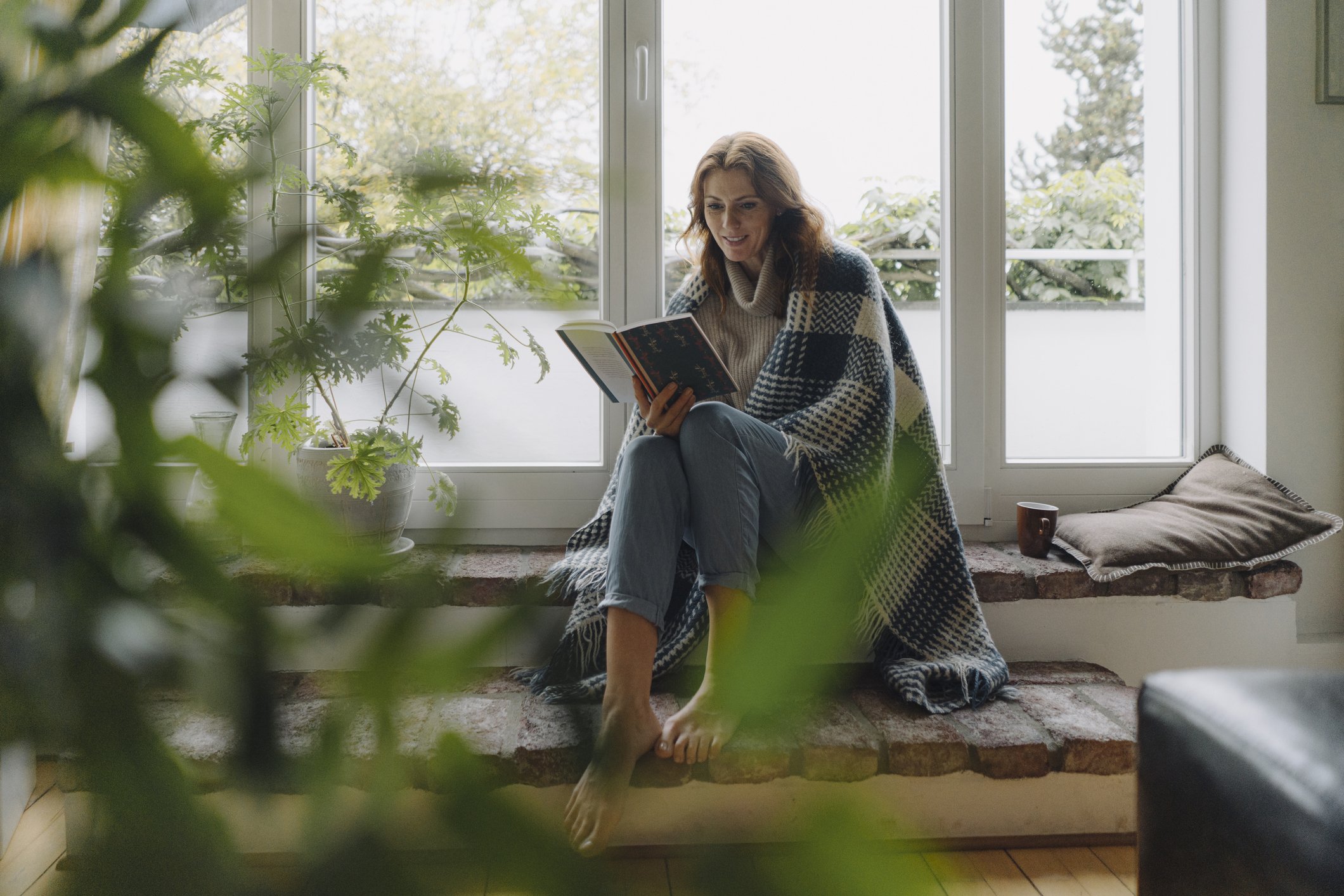 Frau sitzt am Fenster, in Decke gewickelt, und liest ein Buch I Quelle: Getty Images