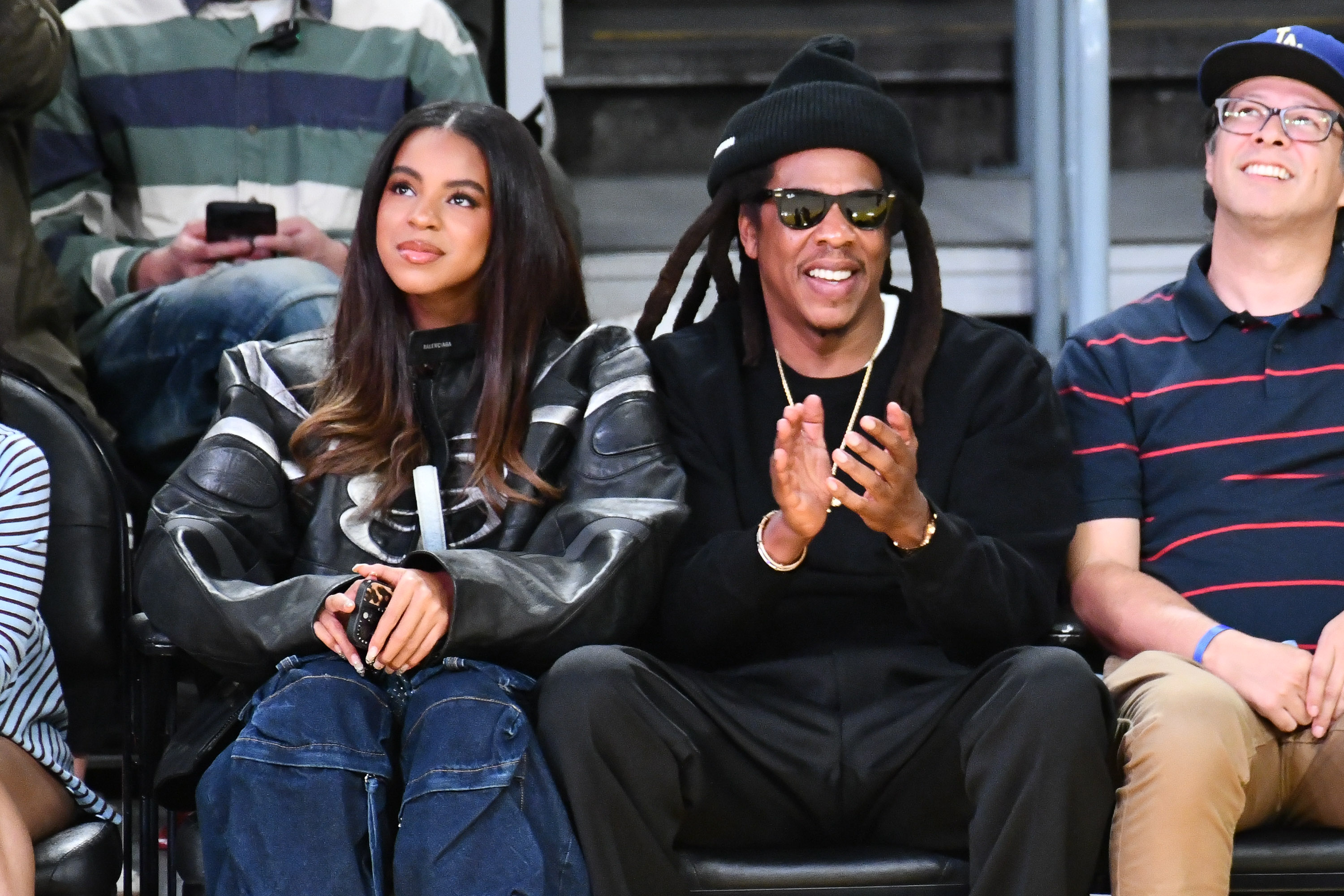Blue Ivy Carter is seen with a relaxed, soft, and slightly amused expression, while her father, Jay-Z, sits beside her with a wide smile as he claps during the game.
