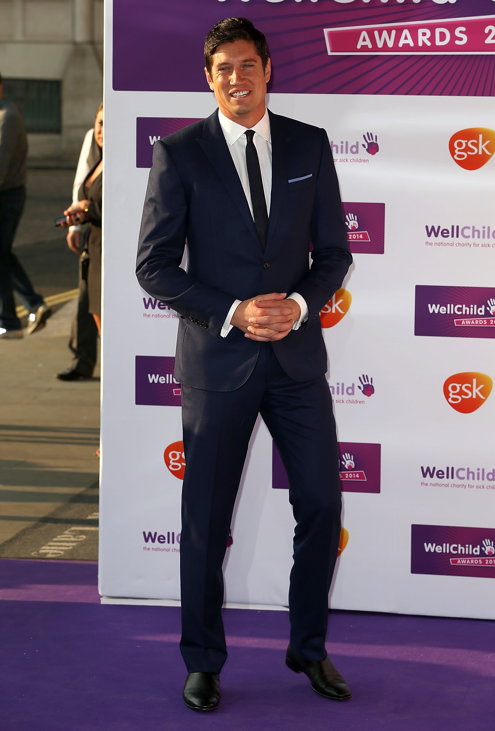 Vernon Kay is dressed in a tailored navy suit with a crisp white shirt and black tie, smiling warmly as he poses on the purple carpet with his hands clasped in front of him.