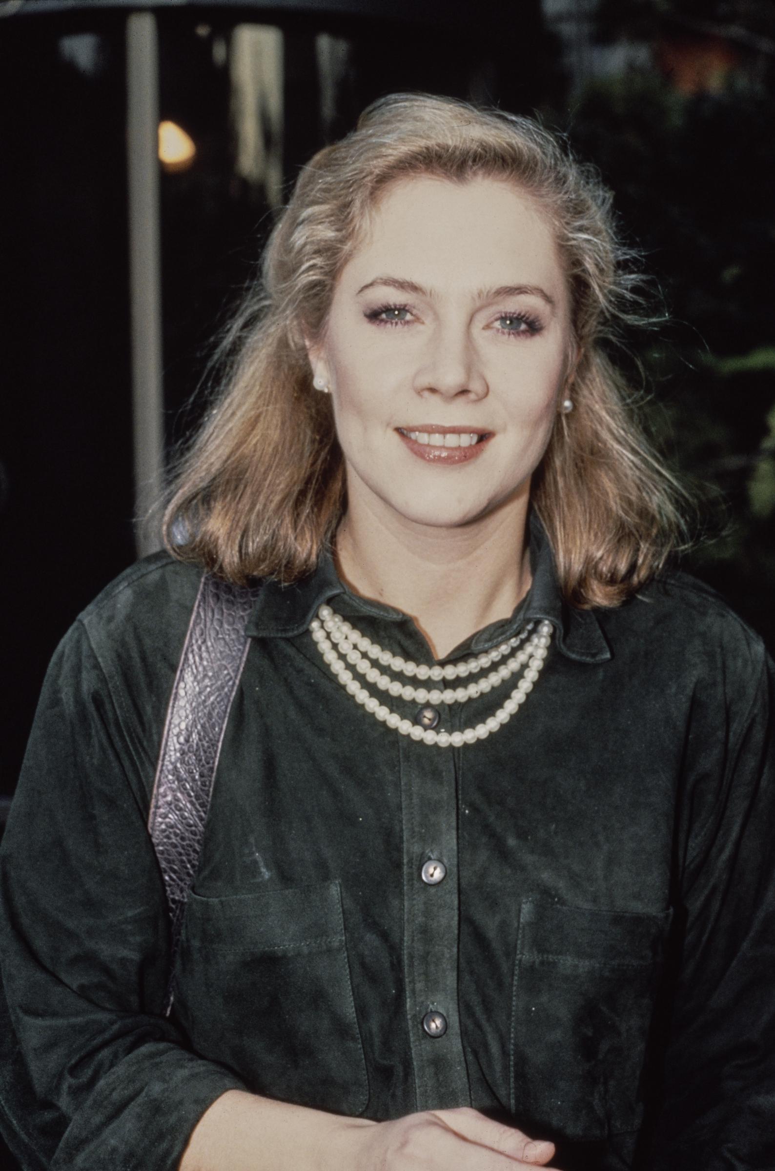 Kathleen Turner is pictured at the rehearsals for the 42nd Golden Globe Awards, looking elegant in a dark green button-up suede shirt paired with a layered pearl necklace. Her softly waved, shoulder-length hair and natural makeup highlight her timeless beauty.