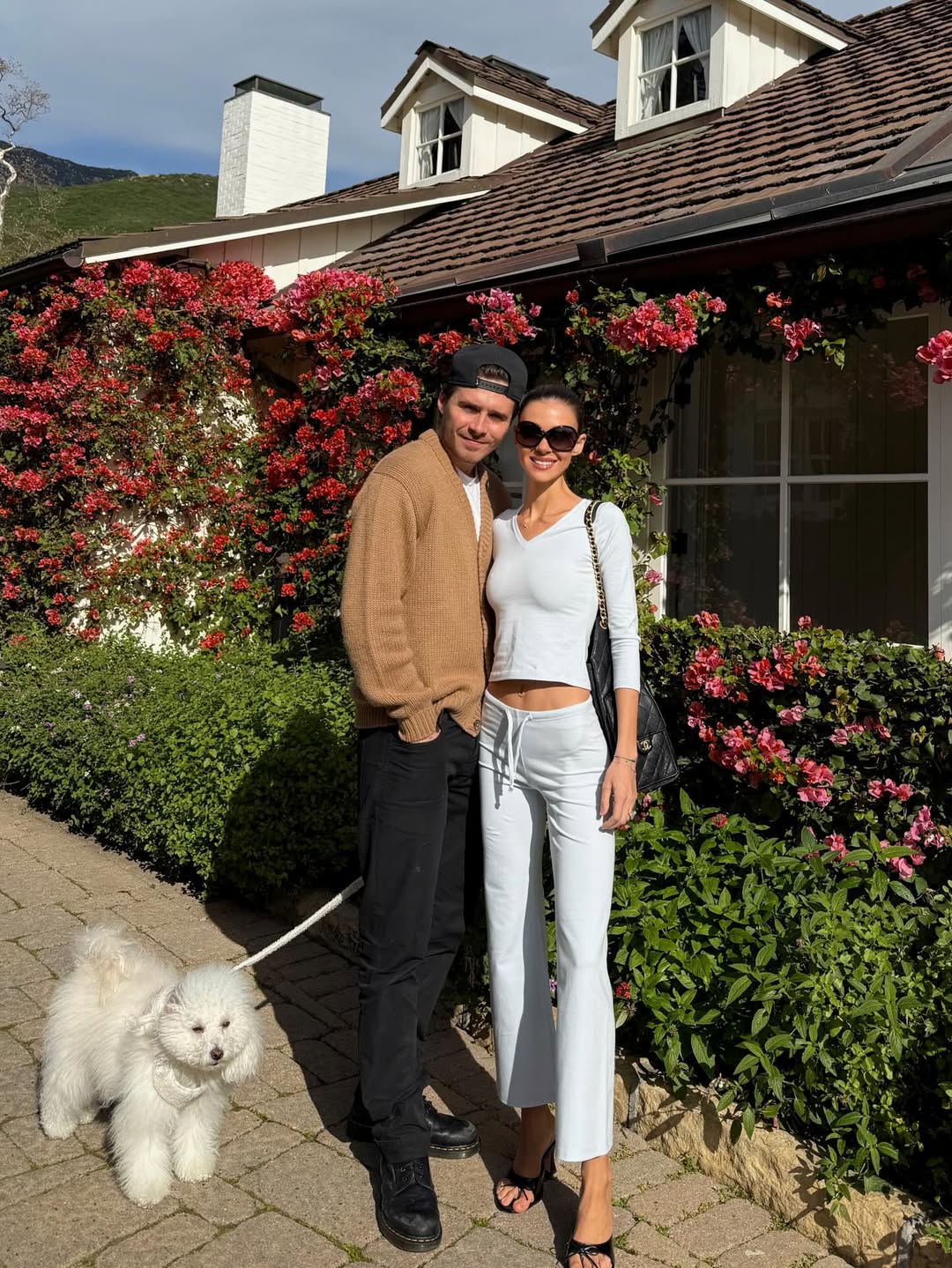Nicola and husband Brooklyn share a sunny outdoor moment with their fluffy pup by their side. Dressed in a soft blue co-ord, Nicola coordinates effortlessly with Brooklyn, who keeps it casual in dark trousers and a neutral cardigan — a relaxed snapshot framed by blooming florals. | Source: Instagram/nicolapeltzbeckham