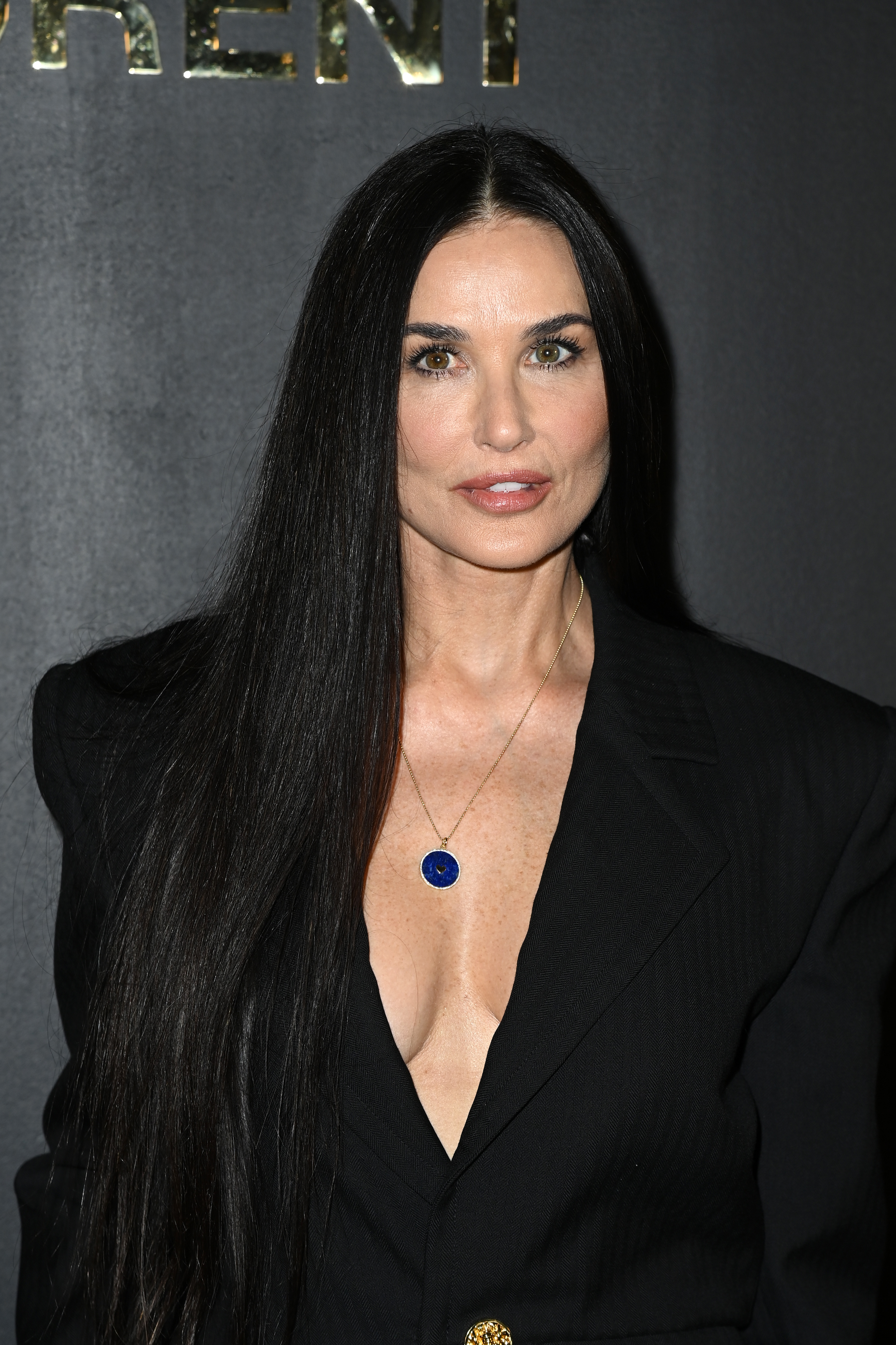 Demi Moore commands attention in a sleek black ensemble, her long, straight hair cascading like silk against the dark backdrop. A deep neckline and a striking blue pendant add a bold touch of contrast and confidence. Her poised expression reflects both strength and timeless sophistication.