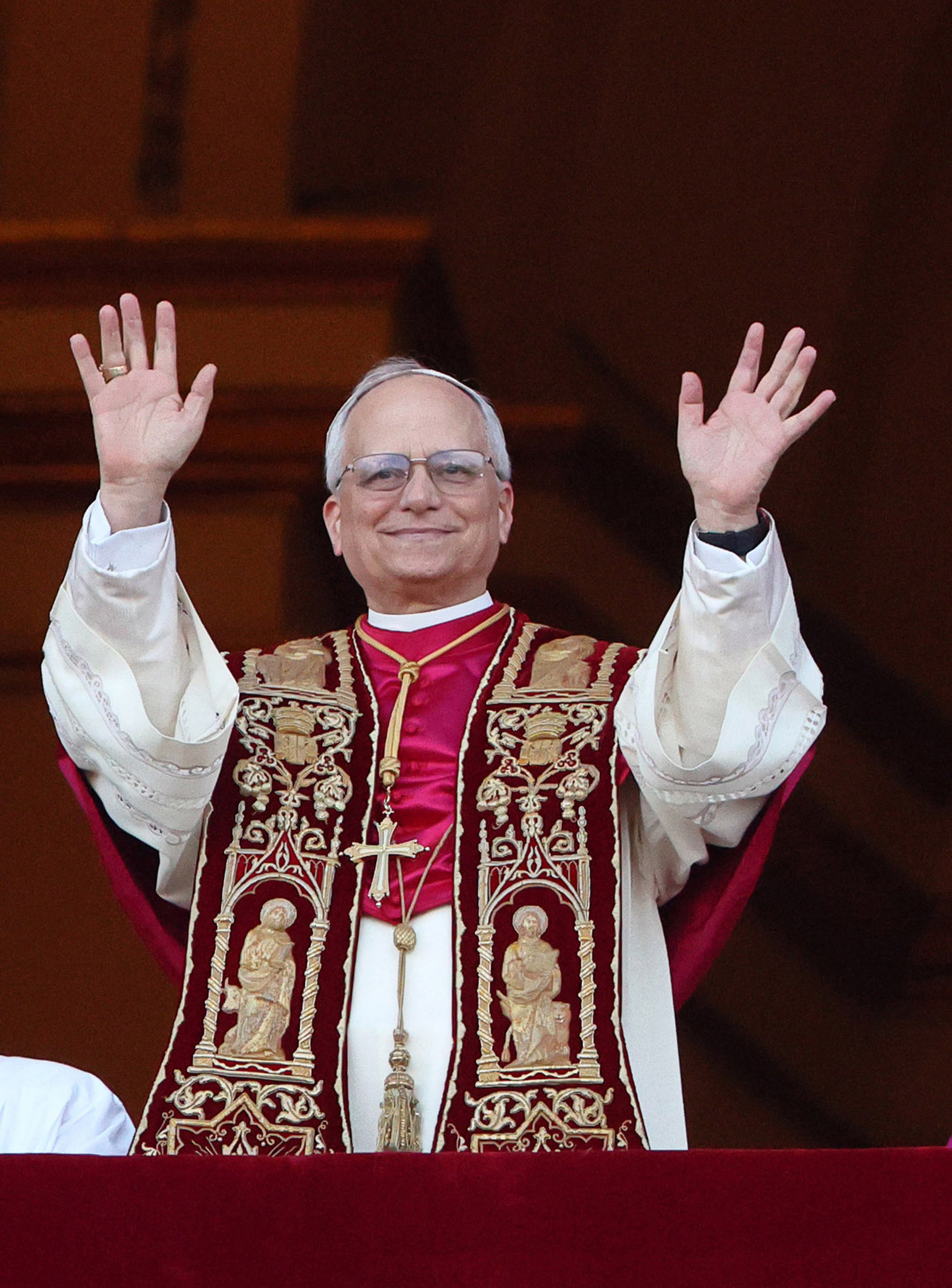 Pope Leo XIV smiles warmly as he raises both hands in greeting, his ornate crimson stole detailed with golden saints catching the light