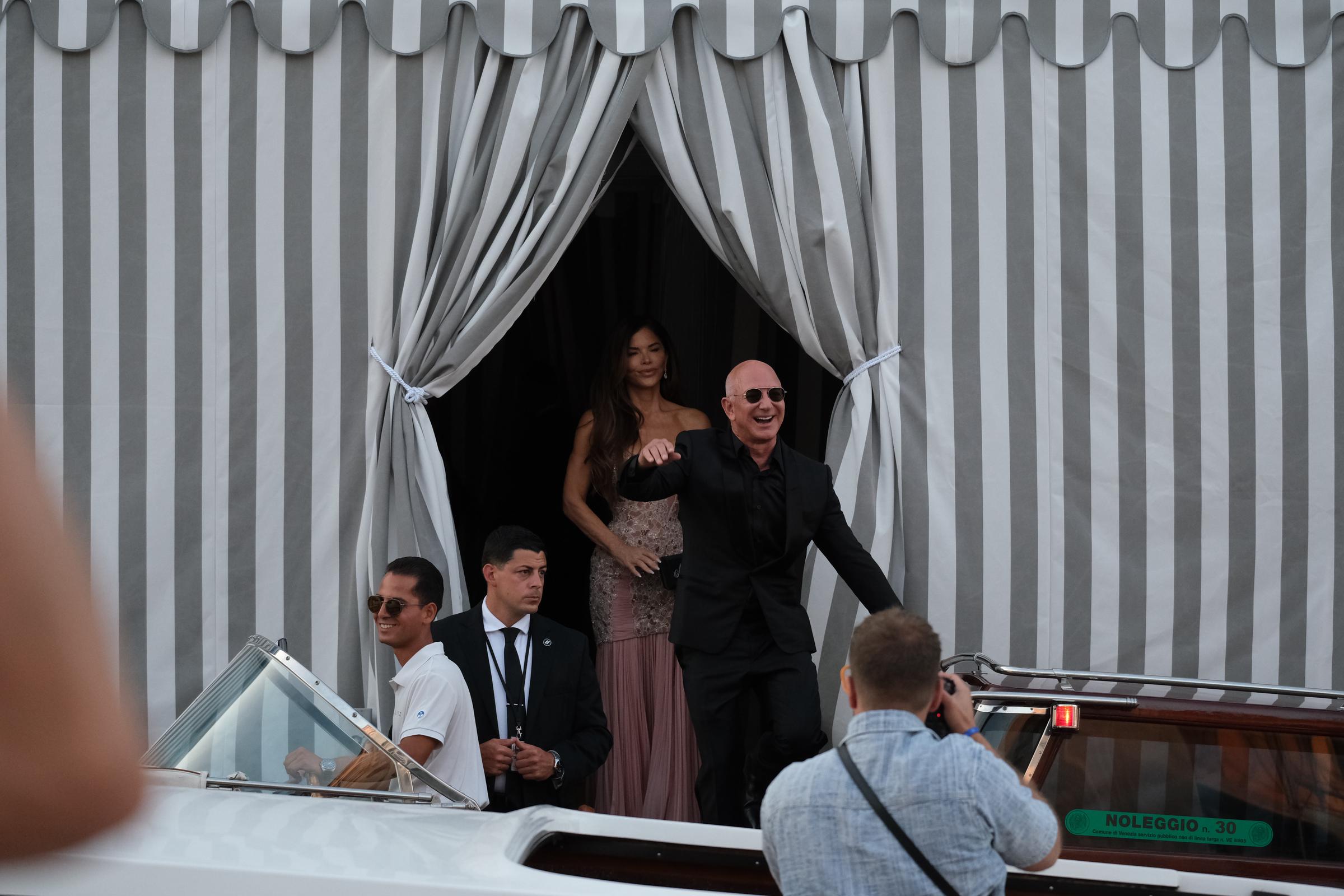 Jeff Bezos beams and waves enthusiastically while stepping onto a water taxi, as Lauren Sánchez follows behind him wearing a blush-toned beaded gown, framed by striped curtains and surrounded by staff and onlookers.