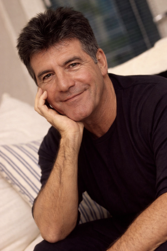 Pictured in a relaxed pose with his chin resting on his hand, he wears a dark top against a softly lit indoor background, salt-and-pepper stubble visible along the jaw. | Source: ChatGPT