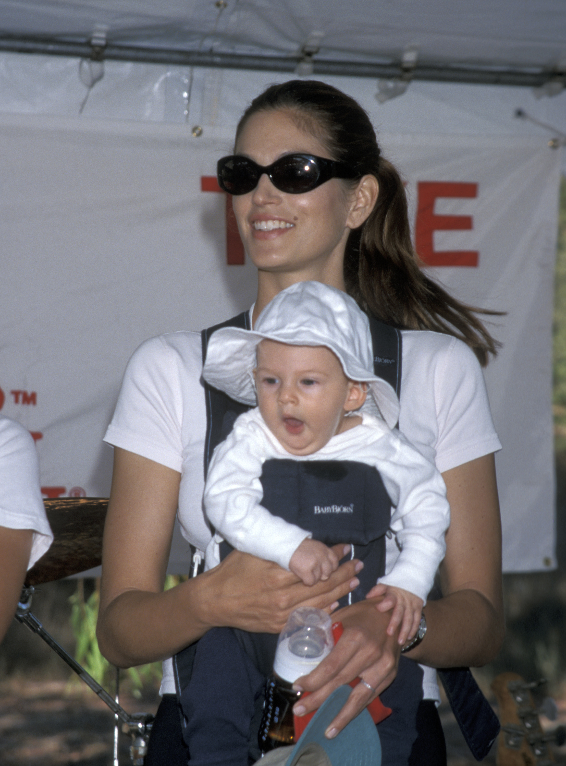 Cindy Crawford carries a baby Presley Gerber at the Take a Hike fundraiser in Malibu in 1999, beaming as she holds him close in a front carrier. Presley, dressed in a floppy white sun hat and matching outfit, yawns softly as he grips his bottle — a quiet reminder of just how tiny and angelic he once was before growing up under the glare of fame.