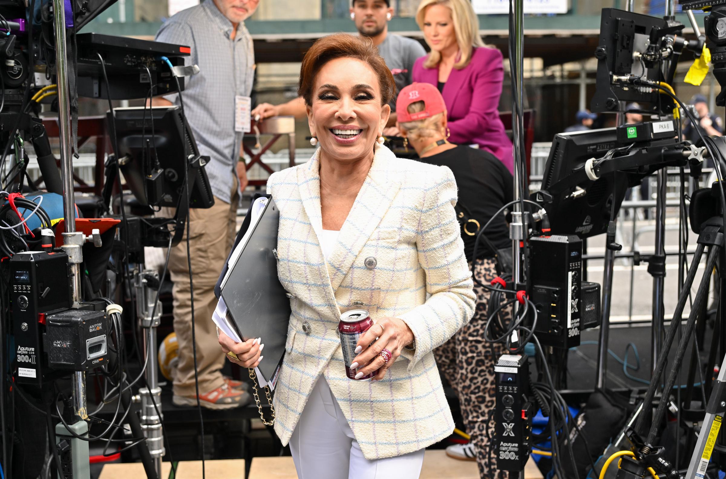 Jeanine Pirro, walks outside the Manhattan Criminal Courthouse. She wears a white plaid blazer over white pants, holding documents and a beverage can, with an upbeat expression amid a busy media setup.