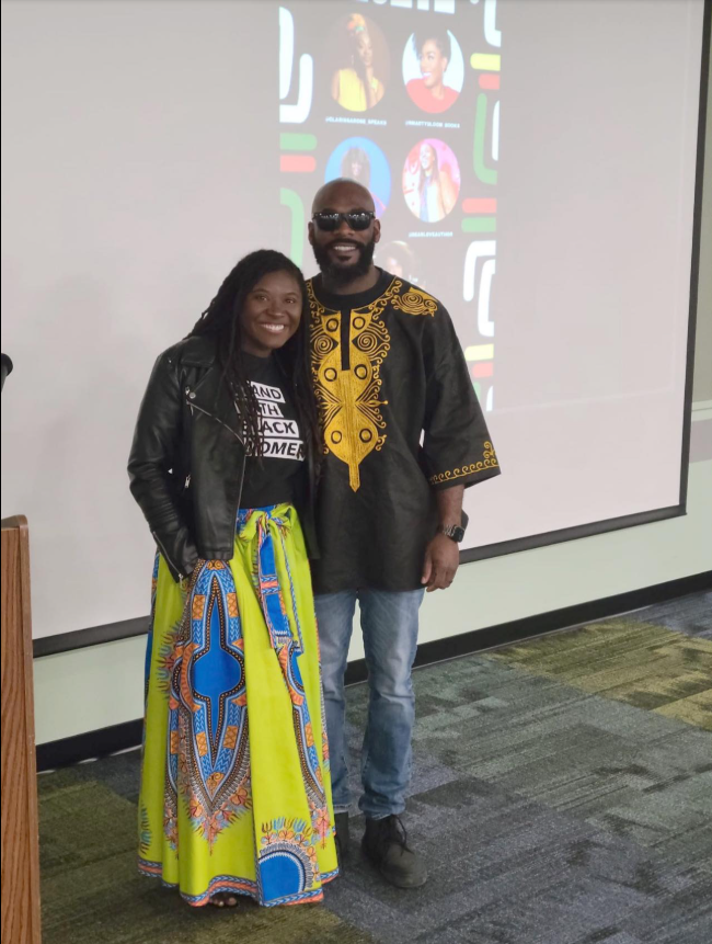 Nancy Metayer Bowen stands beside her husband, Stephen Bowen, smiling warmly in a vibrant patterned skirt and leather jacket, while he wears a black and gold traditional-style top, the couple posing together in front of a presentation screen that hints at a community or cultural event. | Source: Facebook/Nancy Metayer Bowen