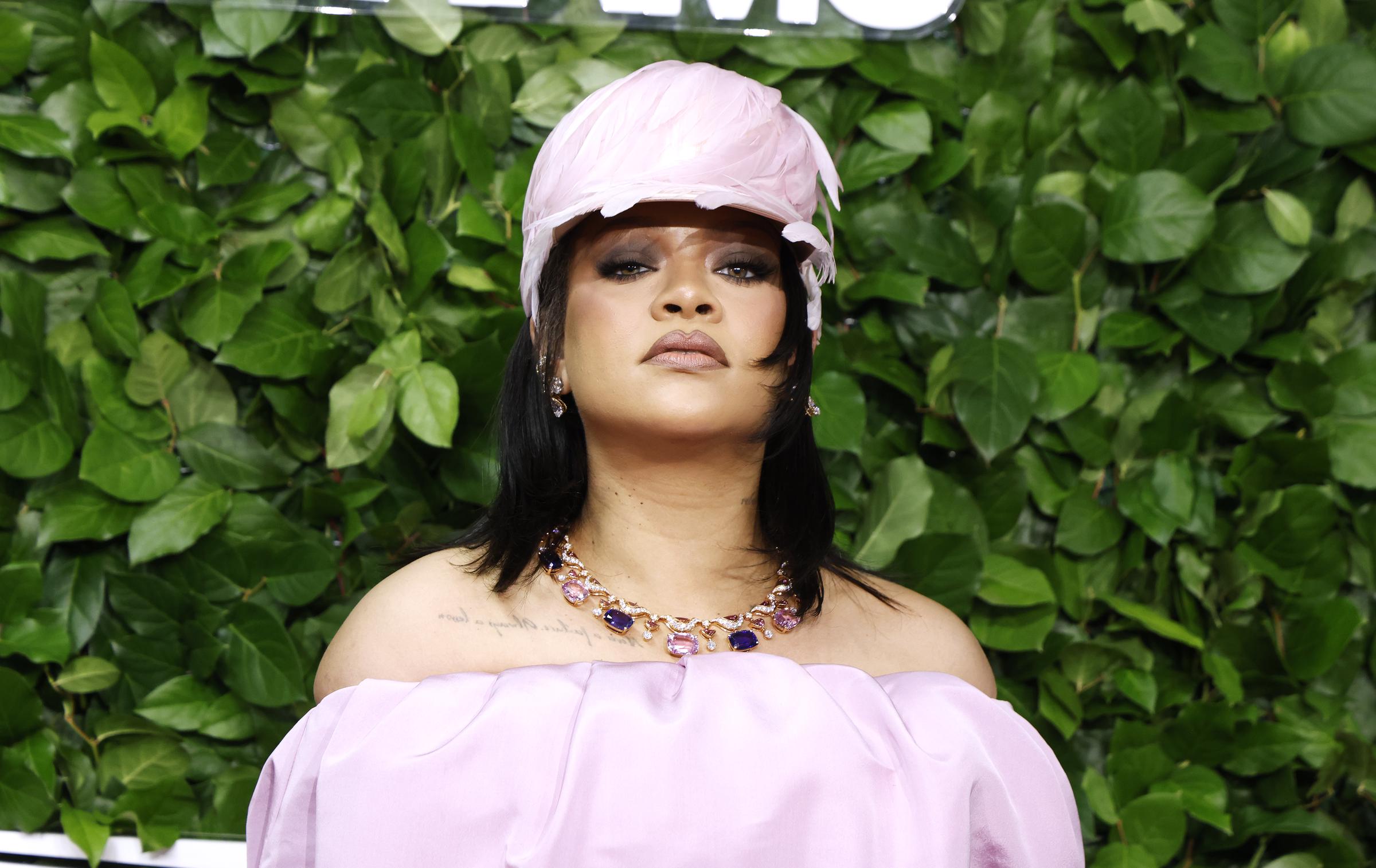 Rihanna poses solo against a lush green backdrop, wearing a pink off-shoulder gown and matching cap, her bold eye makeup and jeweled necklace adding a fierce touch to the look.
