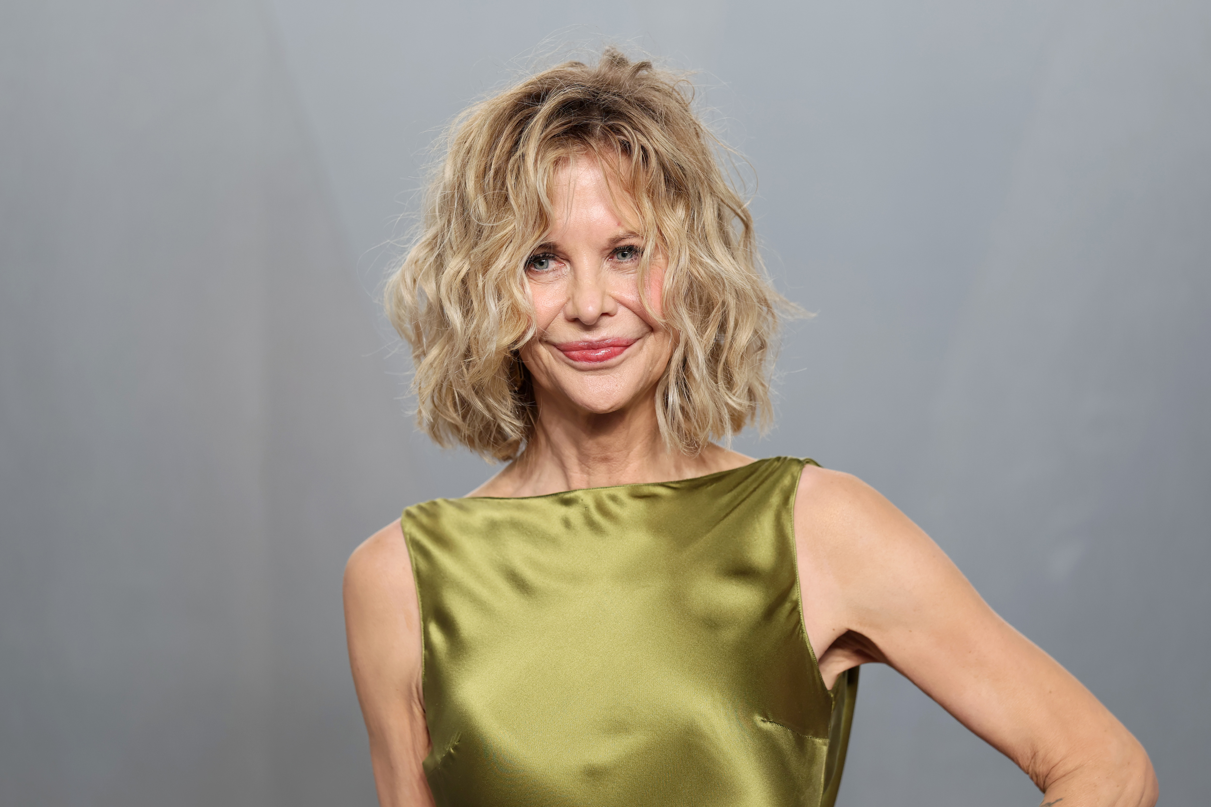 Meg Ryan smiles for photographers at the Vanity Fair Oscar Party, showcasing a sleek olive satin gown and her signature tousled blonde hairstyle.