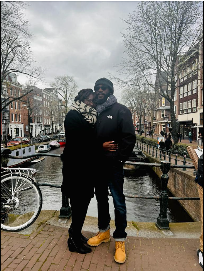Bundled up for cool weather, Nancy Metayer Bowen leans into her husband, Stephen Bowen, as they stand on a canal-side street lined with historic buildings and bicycles, sharing a quiet, affectionate moment during a scenic trip. | Source: Facebook/Nancy Metayer Bowen