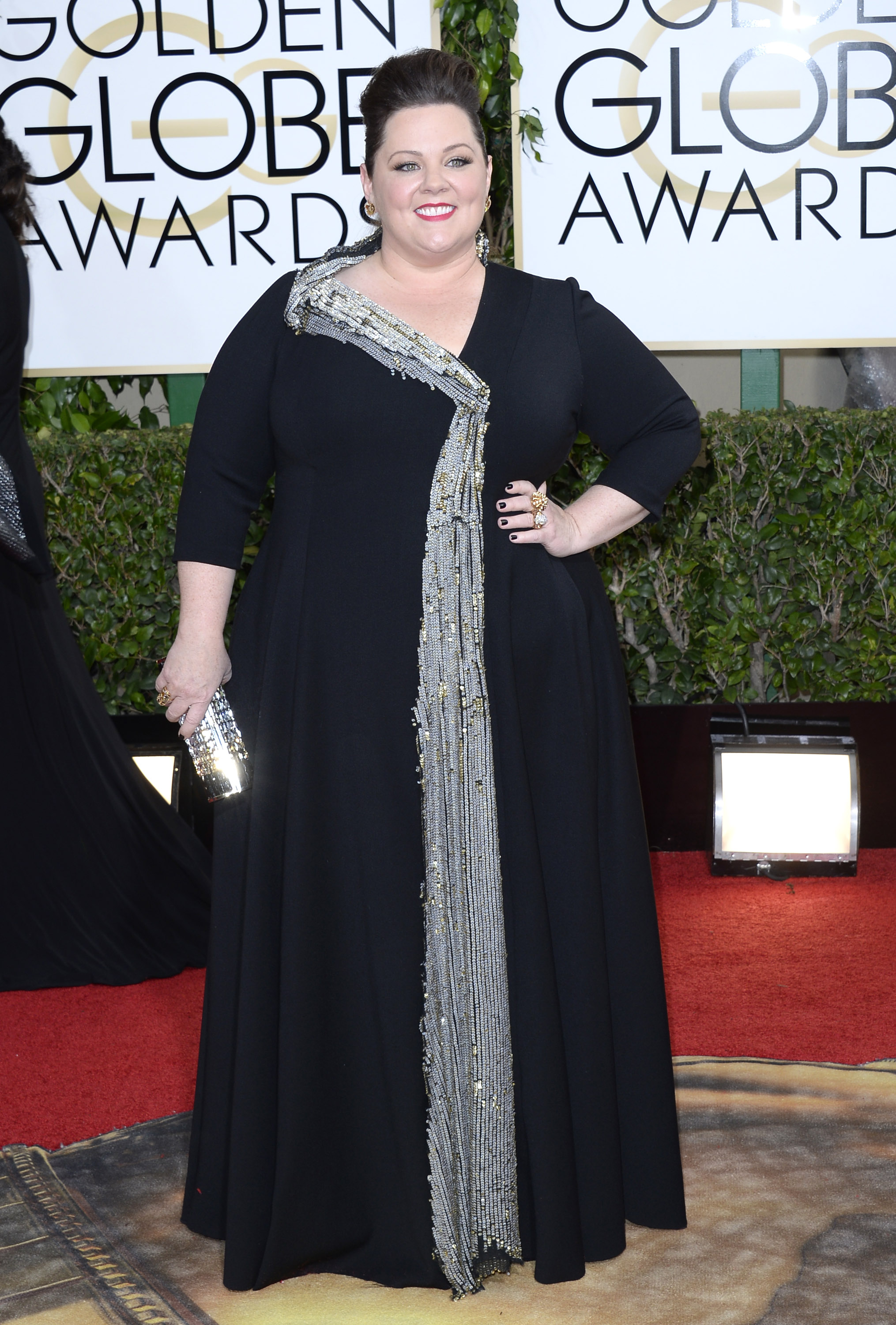 in a striking black gown adorned with a sweeping cascade of shimmering silver embellishments, Melissa McCarthy steps onto the 71st Annual Golden Globe Awards. With a bright smile and confident stance, she brings glamor and poise to the event's spotlight.