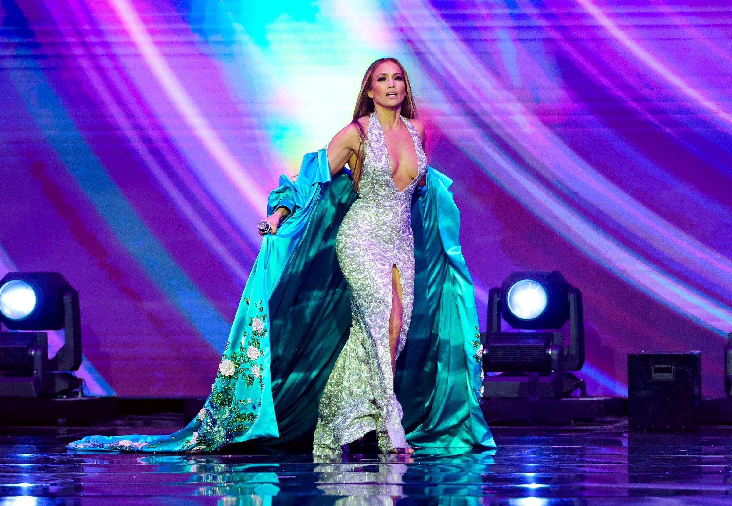 J.Lo strides across the stage with confidence, holding her robe open to reveal the curve-hugging sparkle of her silver gown beneath the dazzling lights.