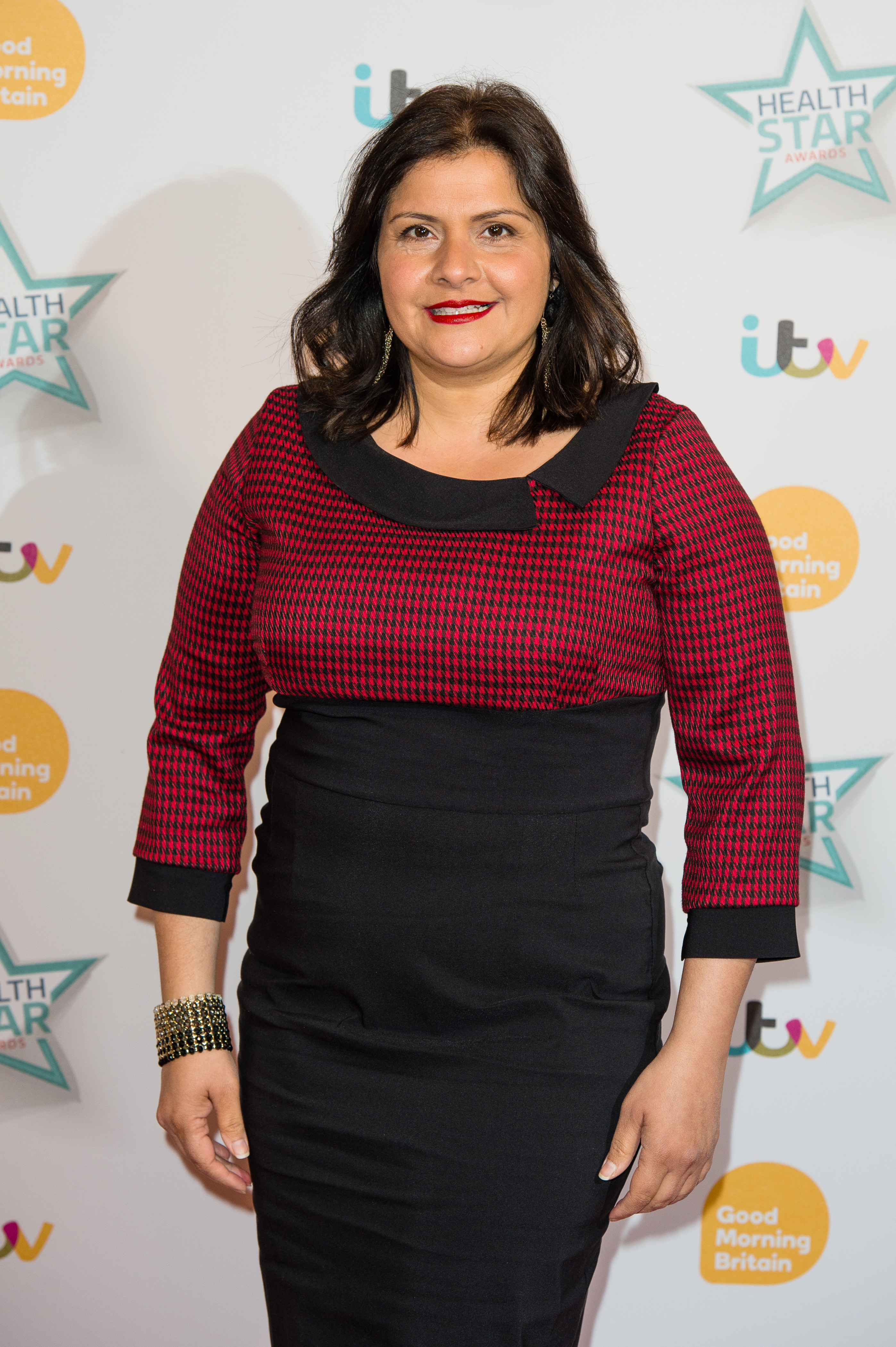 Wadia poses at the Good Morning Britain Health Star Awards at the Hilton on Park Lane in April 2016, wearing a colour-blocked dress combining a red and black houndstooth three-quarter-sleeve top with a fitted black pencil skirt. She finishes the look with bold red lipstick, green drop earrings, and a wide gold studded cuff bracelet.