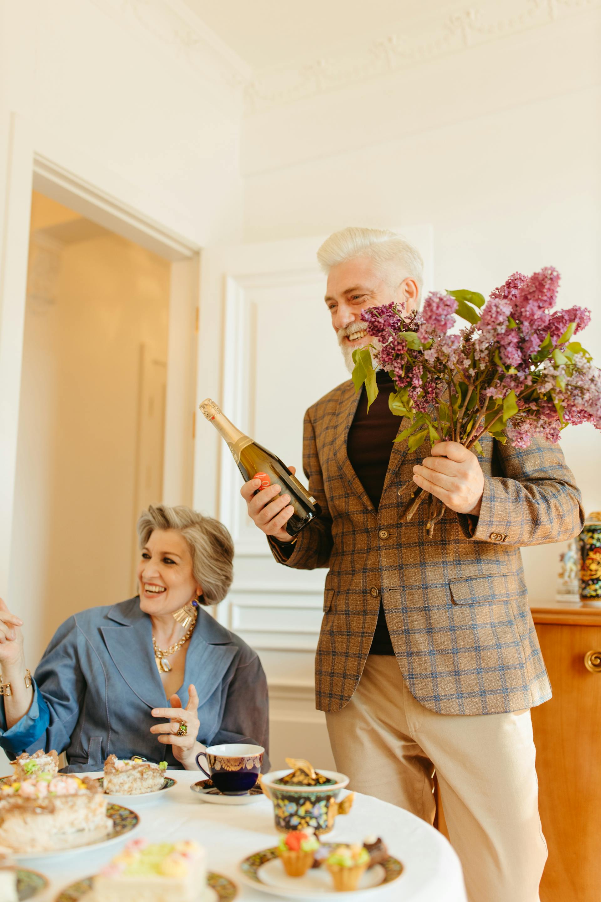 An elderly man celebrating over dinner with a bottle of champagne and flowers while his wife applauds | Source: Pexels