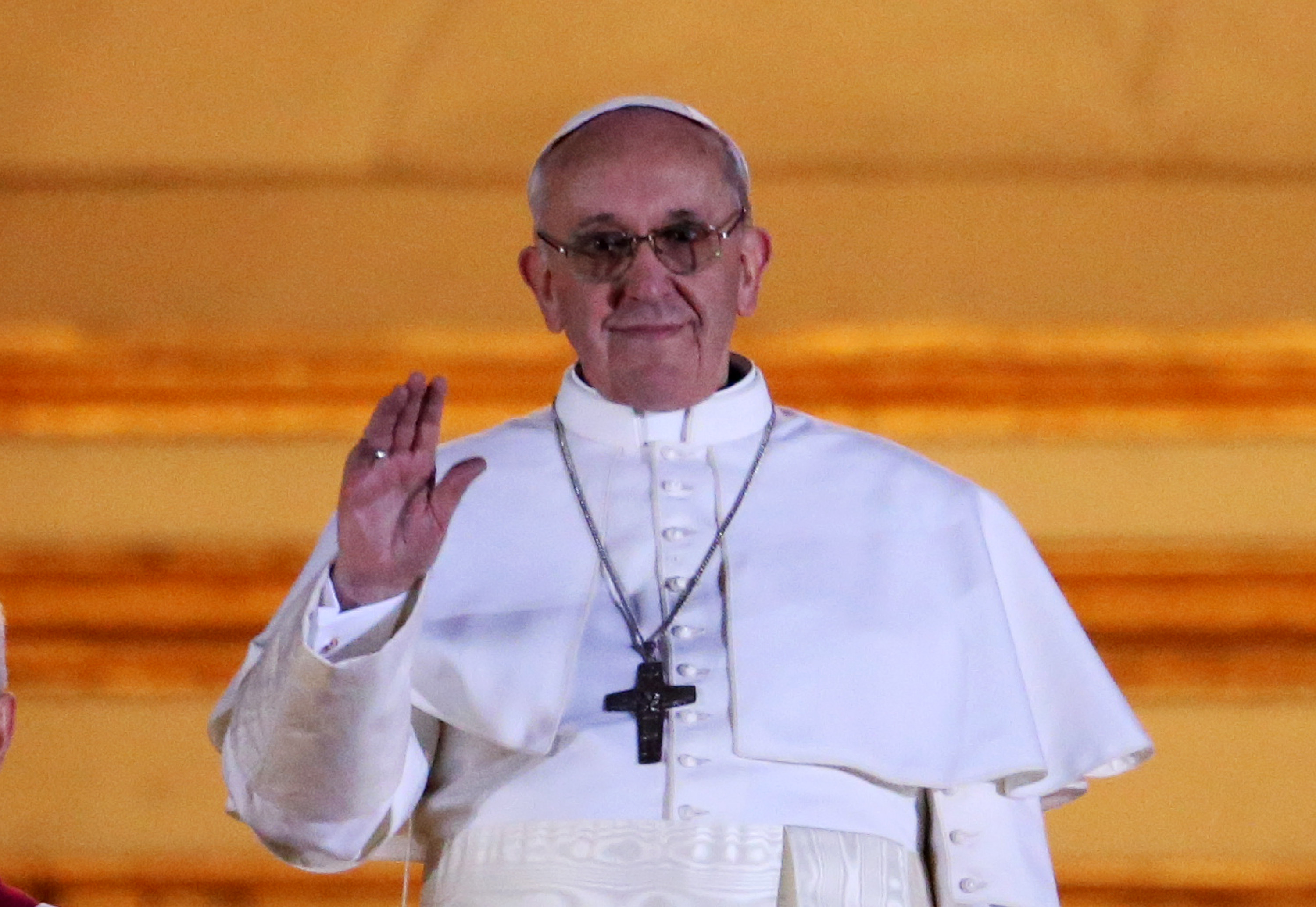 Pope Francis greets the crowd with a gentle wave, dressed in simple white robes and a silver cross, his calm smile framed by soft golden light