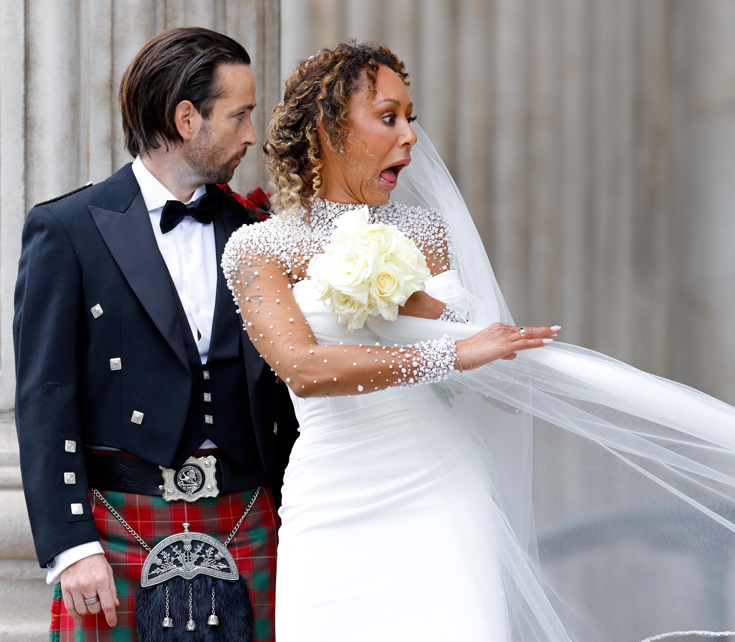 One candid moment shows Mel B caught in a playful struggle with her veil. With a wide-eyed, surprised expression, she reaches up to catch it as it shifts in the breeze—adding a touch of humor and spontaneity to the day’s formal elegance.