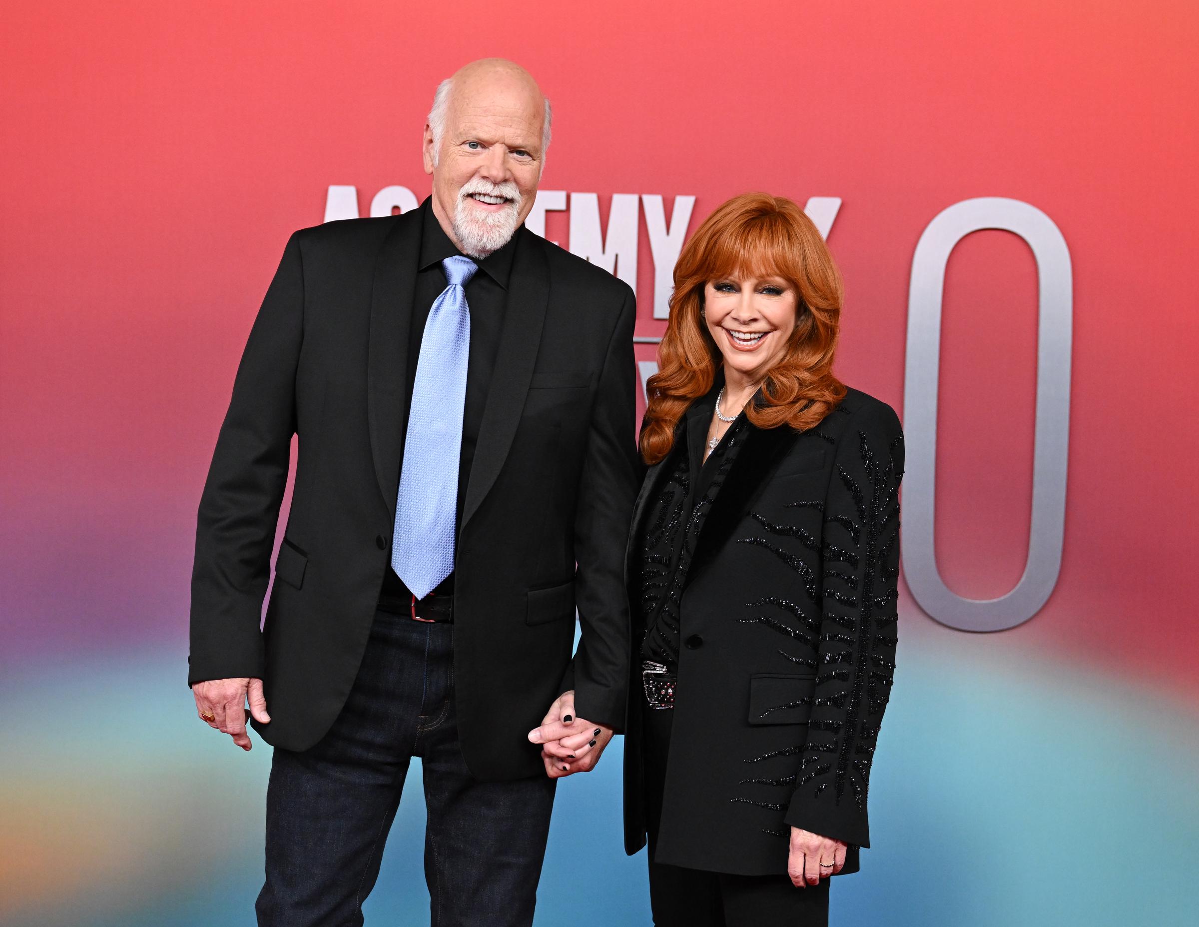 Reba McEntire beams in a black embellished blazer and satin blouse, standing hand-in-hand with Rex Linn, who sports a light blue tie over a black shirt and jeans