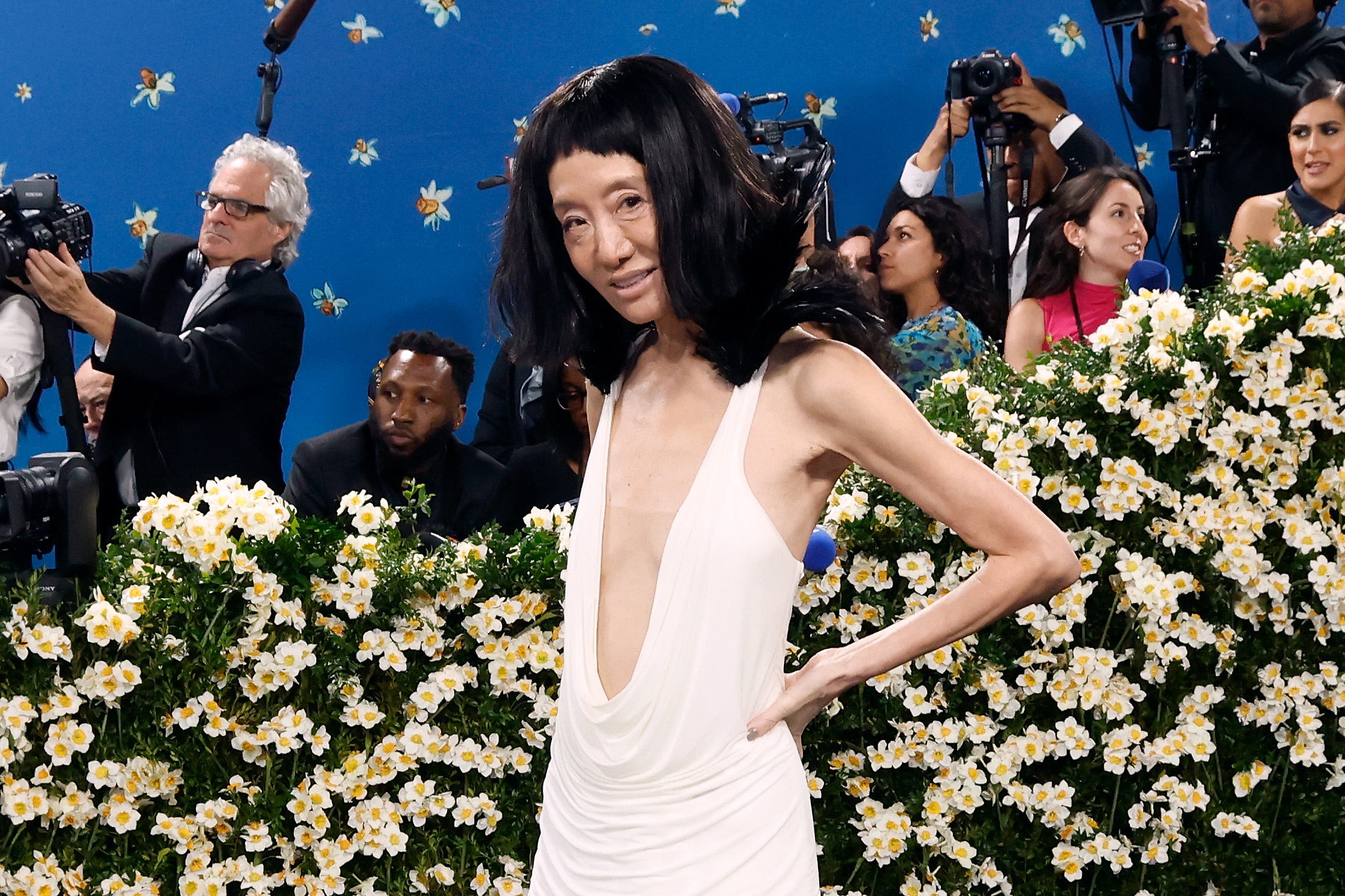 Striking a confident pose, Vera Wang stands against a field of white blossoms, highlighting the plunging neckline and feathered shoulders of her custom gown.