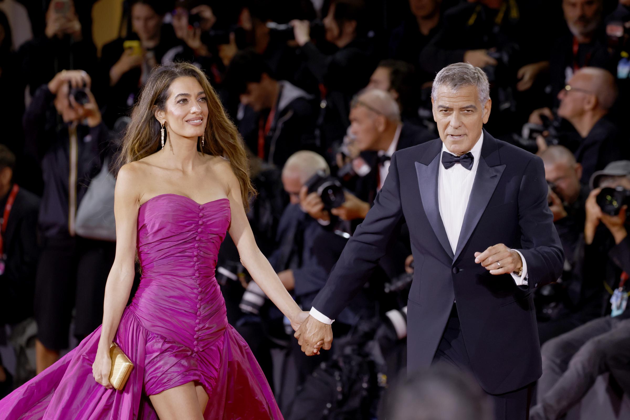 Hand in hand, the star couple strides down the red carpet. Amal's fuchsia gown flows behind her as she holds a gold clutch, while George, in a sharp tuxedo and bow tie, leads with a focused expression through the crowd of flashing cameras.