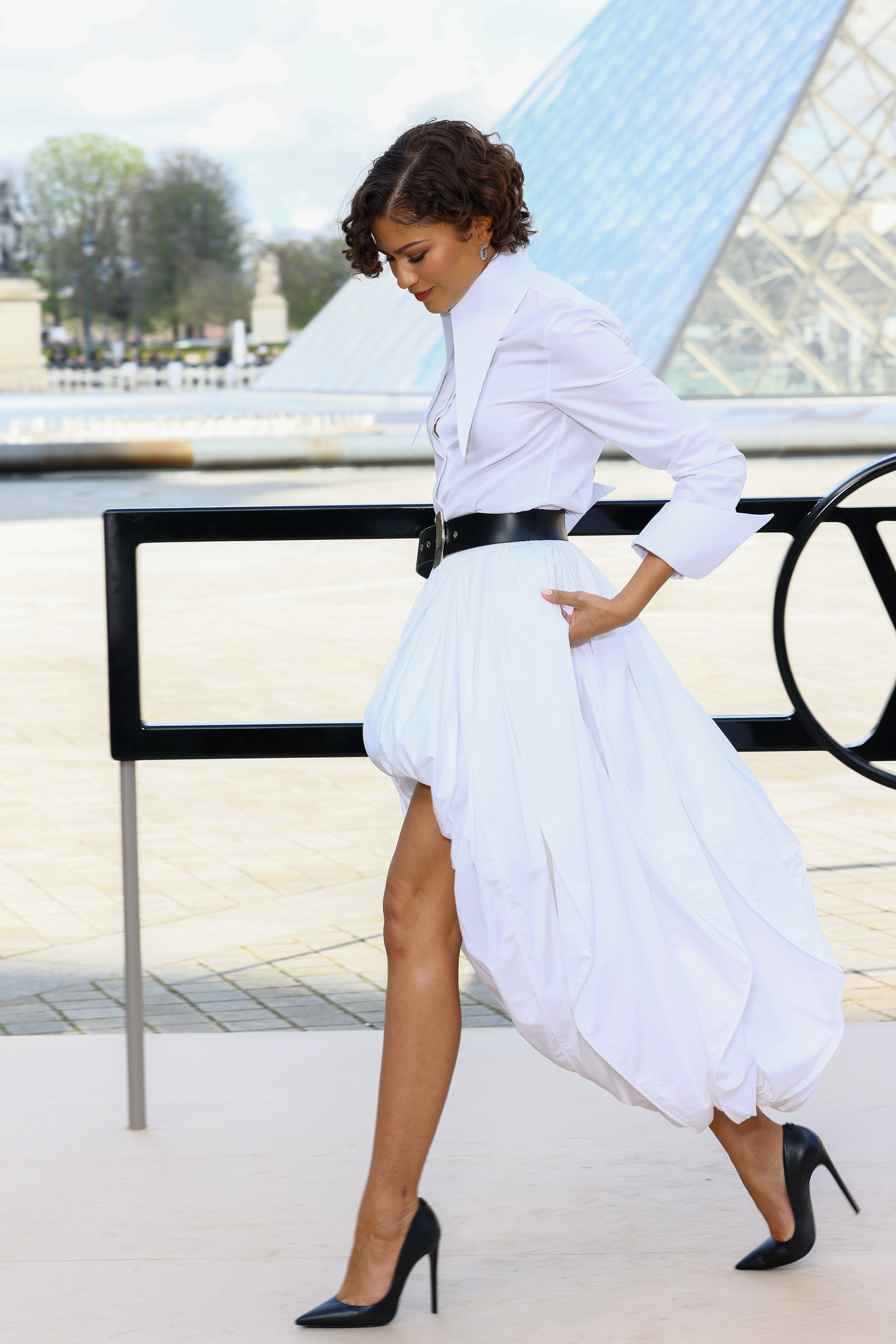 Zendaya walks past a black railing with the Louvre’s glass pyramid visible behind her at the Louis Vuitton Fall/Winter show during Paris Fashion Week in Paris on March 10, 2026. | Source: Getty Images