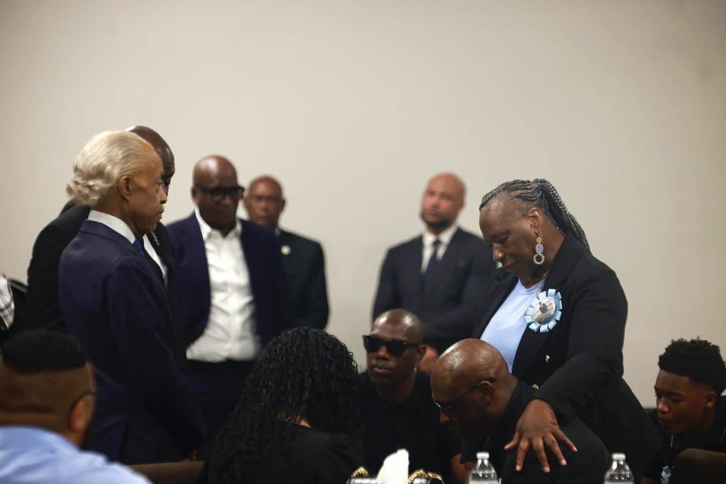 A supporter wearing a memorial pin places a comforting hand on a grieving family member as Rev. Al Sharpton and attendees bow their heads beside Christine Wonsley. | Source: Facebok/realsharpton