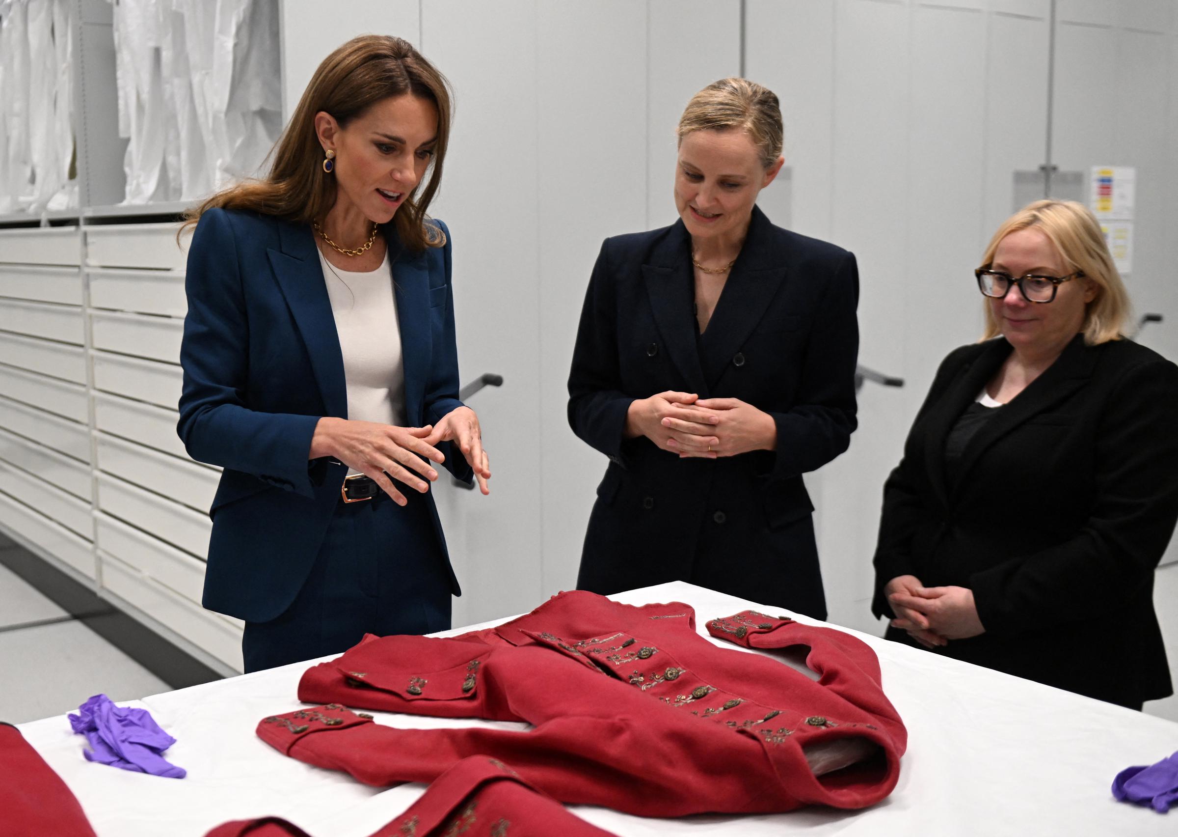 Princess Catherine examines a historic red riding habit alongside curators Oriole Cullen and Kate Parsons. Focused and engaged, she gestures thoughtfully while discussing the garment’s fine detailing and textile preservation.