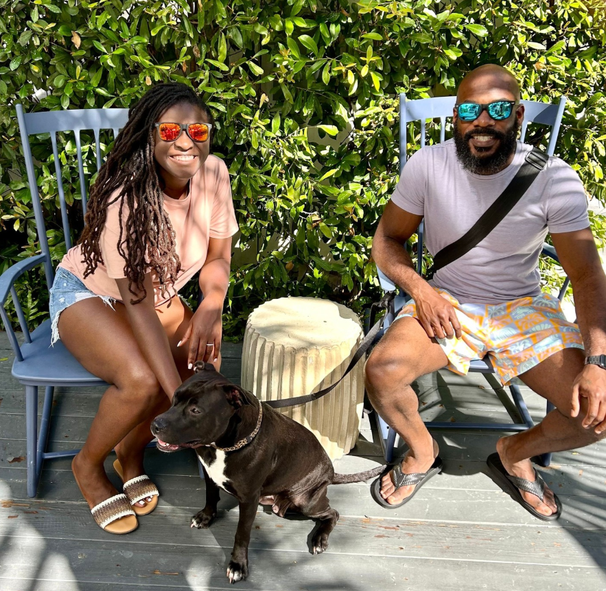 Nancy Metayer Bowen and her husband, Stephen Bowen, sit outdoors on wooden chairs in casual summer outfits, both wearing sunglasses as they smile beside their dog, surrounded by lush greenery in a relaxed daytime setting. | Source: Instagram/nancymetayerbowen