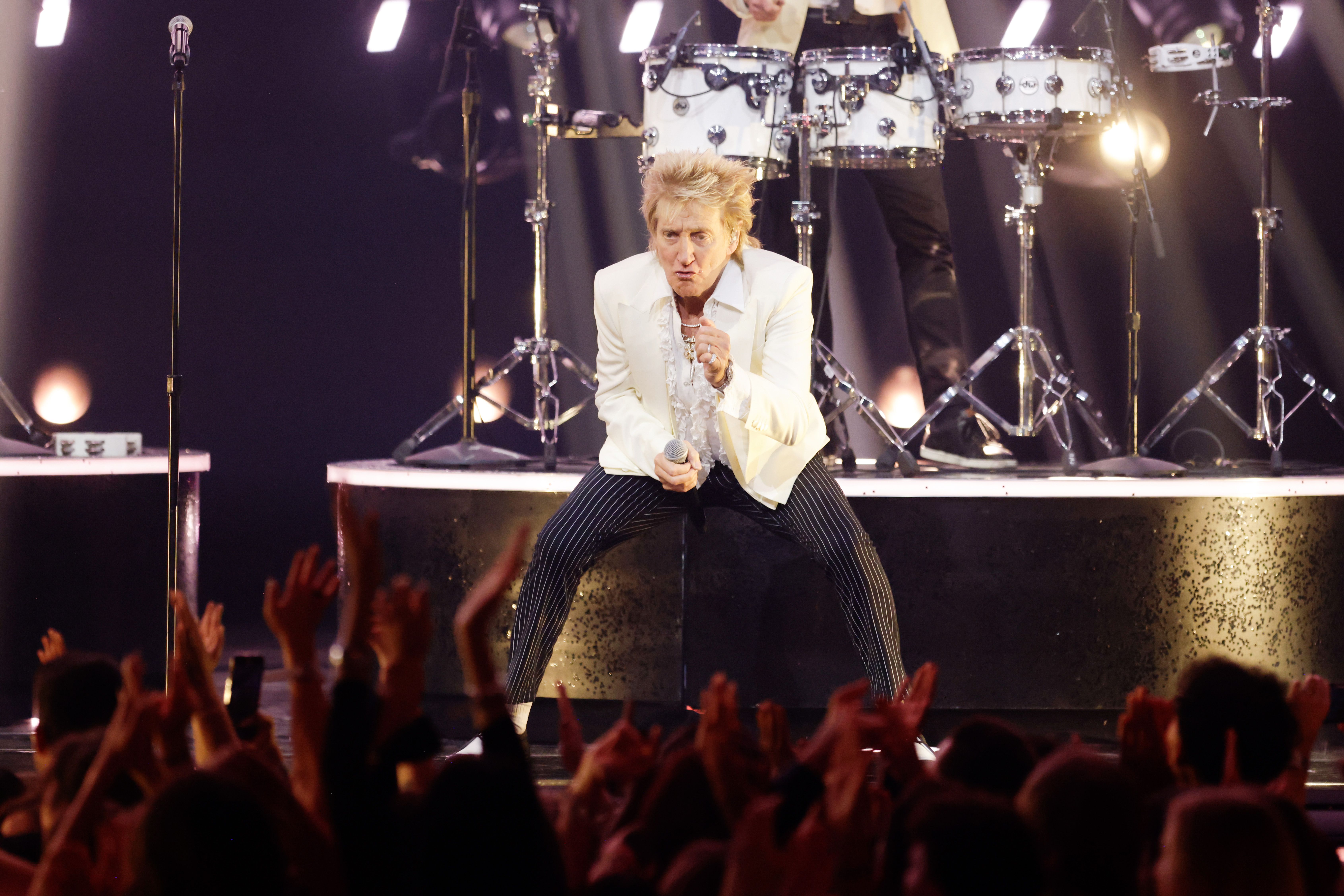 Rod commands the stage in a low, powerful stance, gripping the mic and locking eyes with the crowd. Dressed in a cream jacket, ruffled shirt, and pinstriped pants, he delivers with intensity as fans reach toward him, energized by his every move.