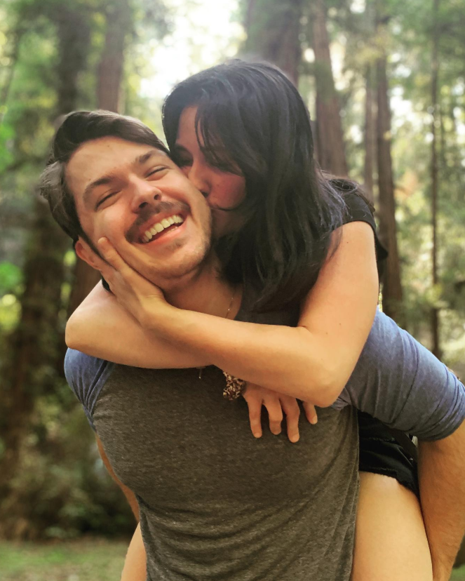 Dakota Norris laughs as he carries his partner on his back, her arms wrapped around him while she kisses his cheek. Set against a forest backdrop, the moment feels playful, affectionate, and full of carefree joy. | Source: Instagram/d.ak.ot.a