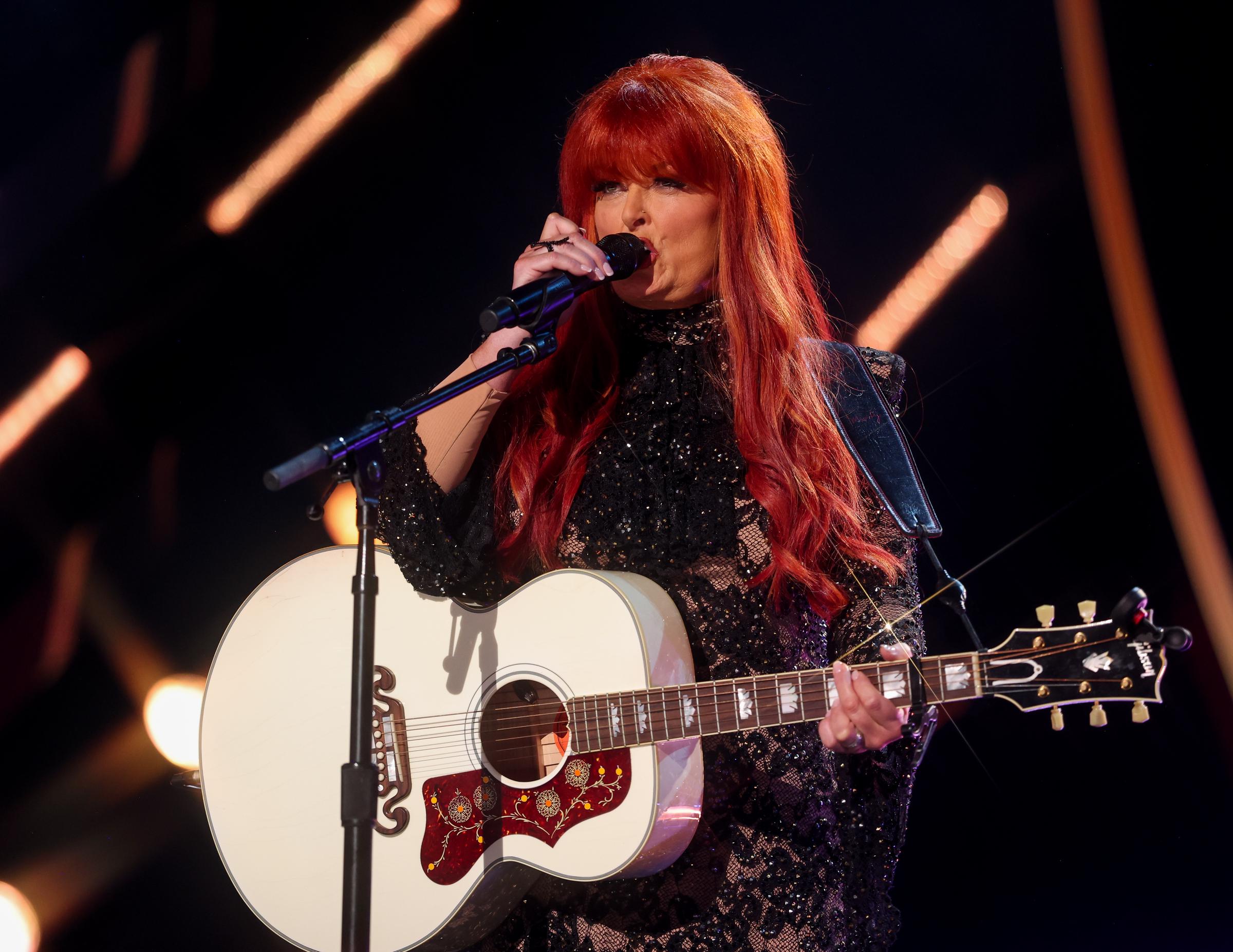 Wynonna Judd plays guitar and sings onstage at the 60th Academy of Country Music Awards. Her intricate black outfit and bright red hair added visual flair to a performance that celebrated her enduring country legacy.