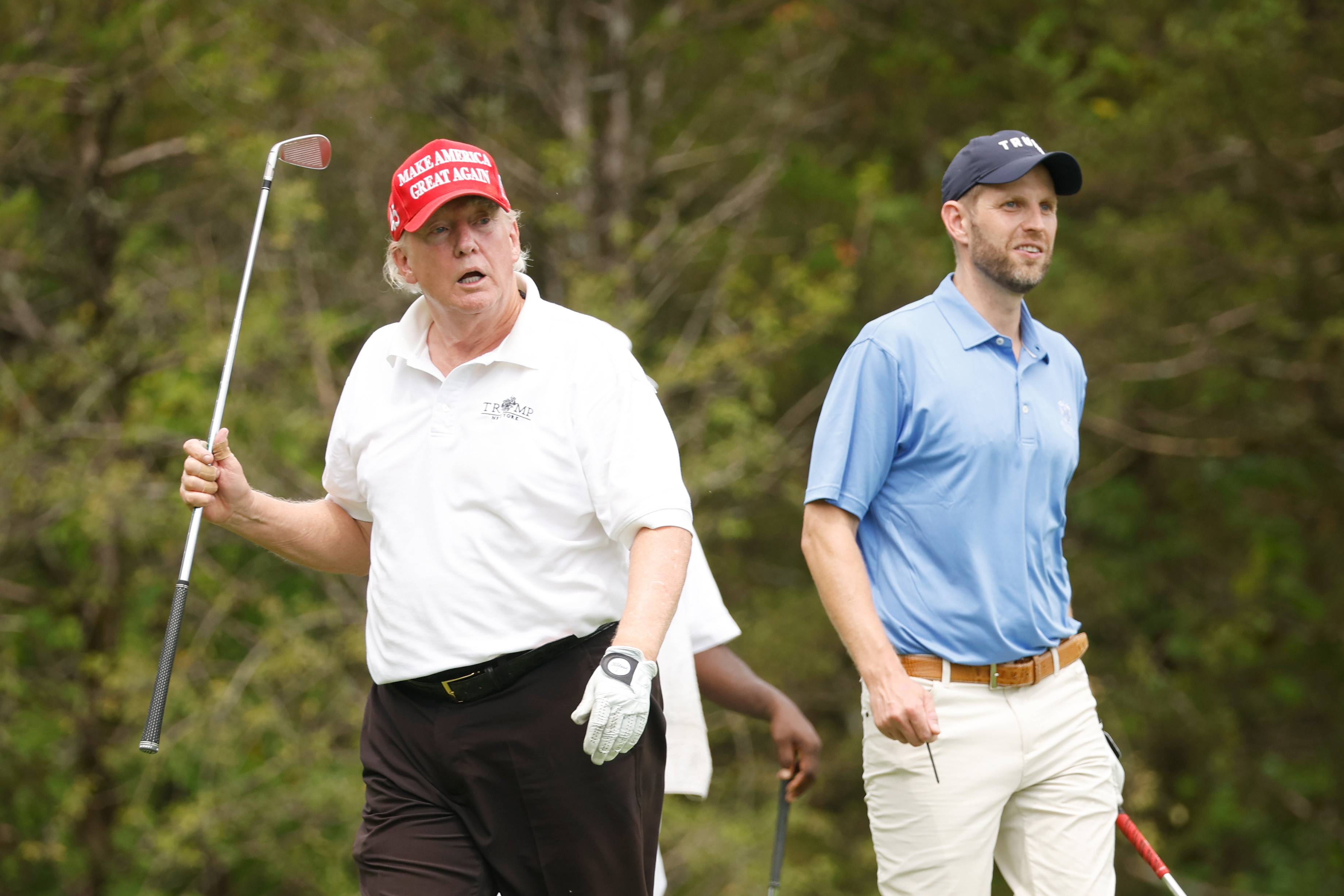 Donald Trump walks alongside his son, Eric Trump, during the tournament, holding a club mid-air. His face shows visible redness around the cheeks and nose, while the rest of his skin looks lighter.