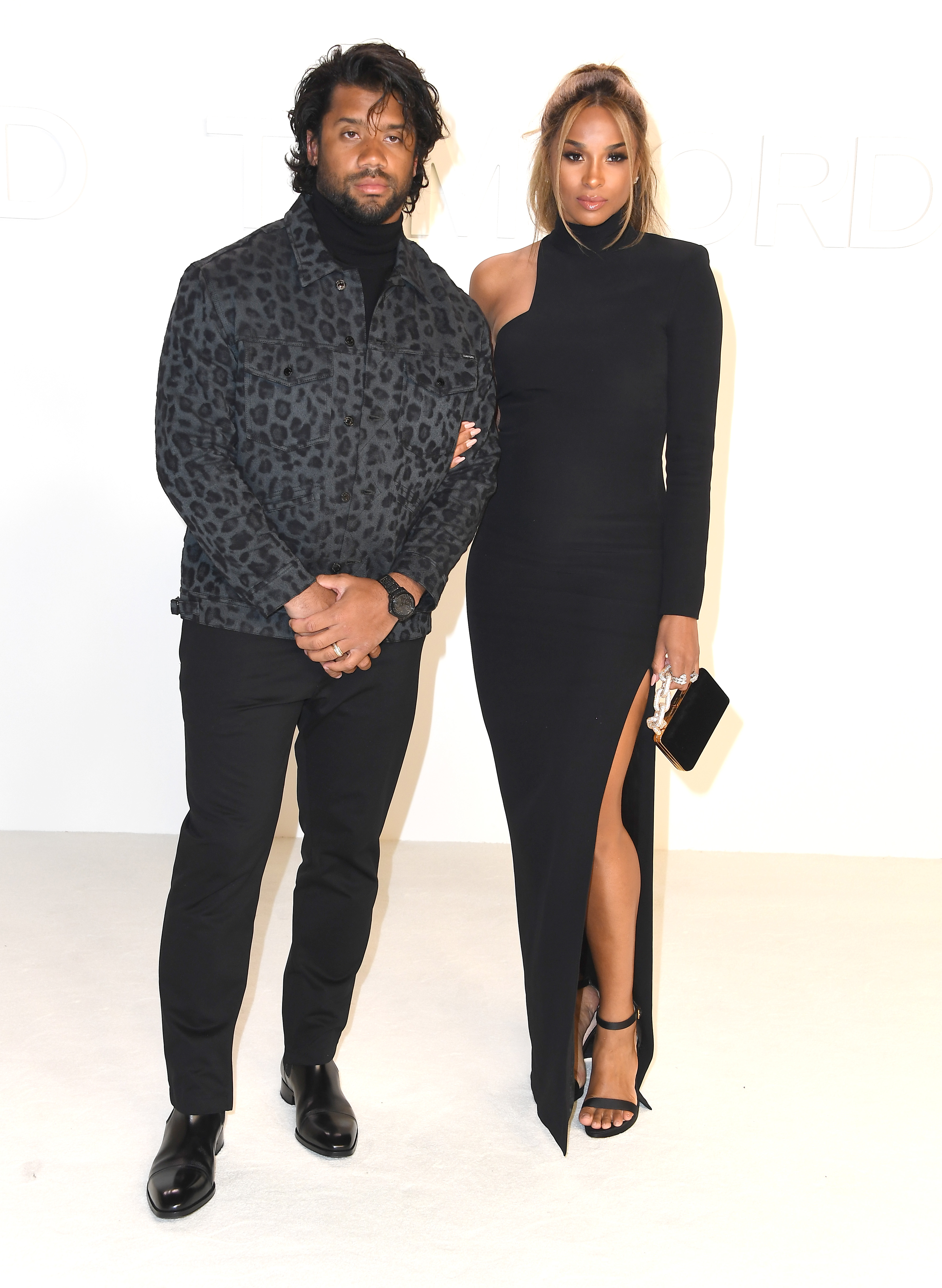 Russell Wilson and Ciara stand side by side in modern, fashion-forward outfits that reflect a sleek and contemporary style. Both maintain composed, confident expressions, giving the moment a clean and effortlessly stylish feel.