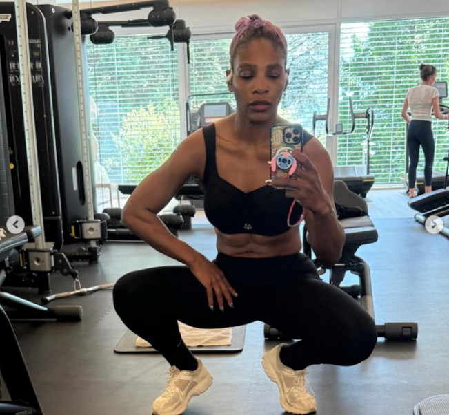 Serena Williams crouched low in front of the gym mirror, holding her phone with one hand while her other rested on her thigh. She wore a black sports bra, matching leggings, white sneakers, and a light pink head wrap.