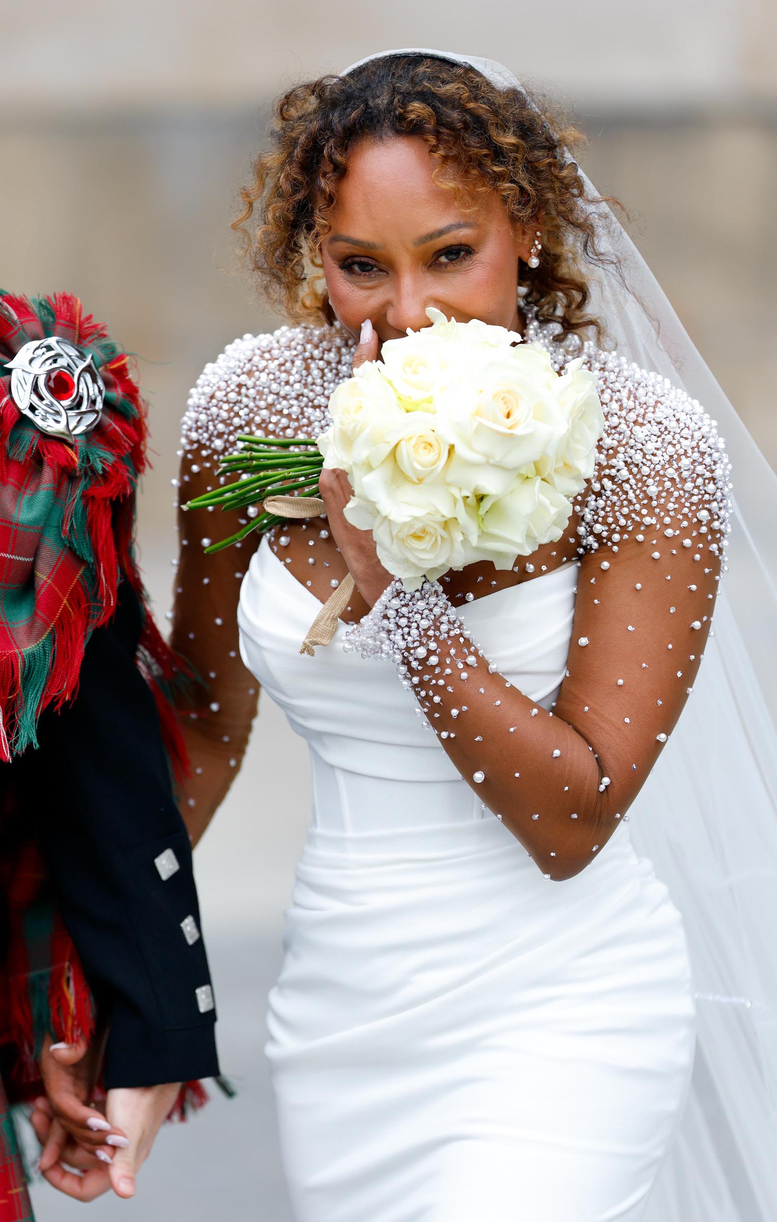 Mel B flashes a warm smile as she holds her bouquet close to her face. The soft petals frame her features, capturing a moment of quiet joy and bridal glow in front of the camera.