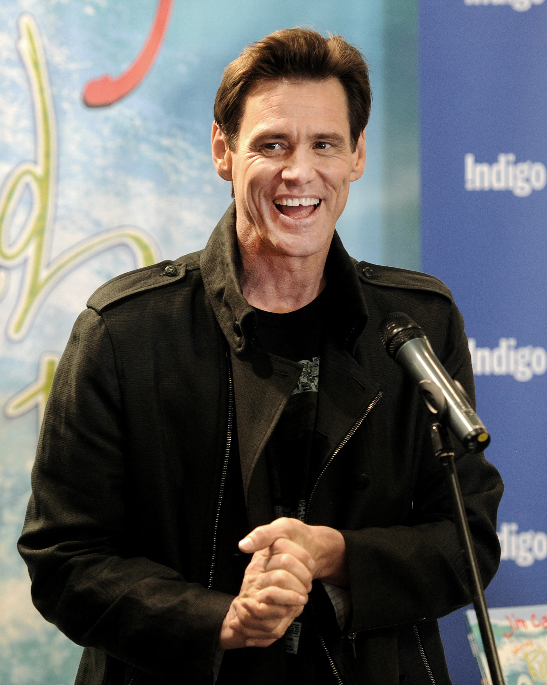 Jim Carrey speaks enthusiastically during a signing event for his children’s book "How Roland Rolls" at Indigo in Yorkdale Mall. Dressed in a black jacket, he smiles broadly while engaging the audience with his signature warmth and humor. The colorful backdrop and microphone highlight the lively, joyful atmosphere of the occasion.