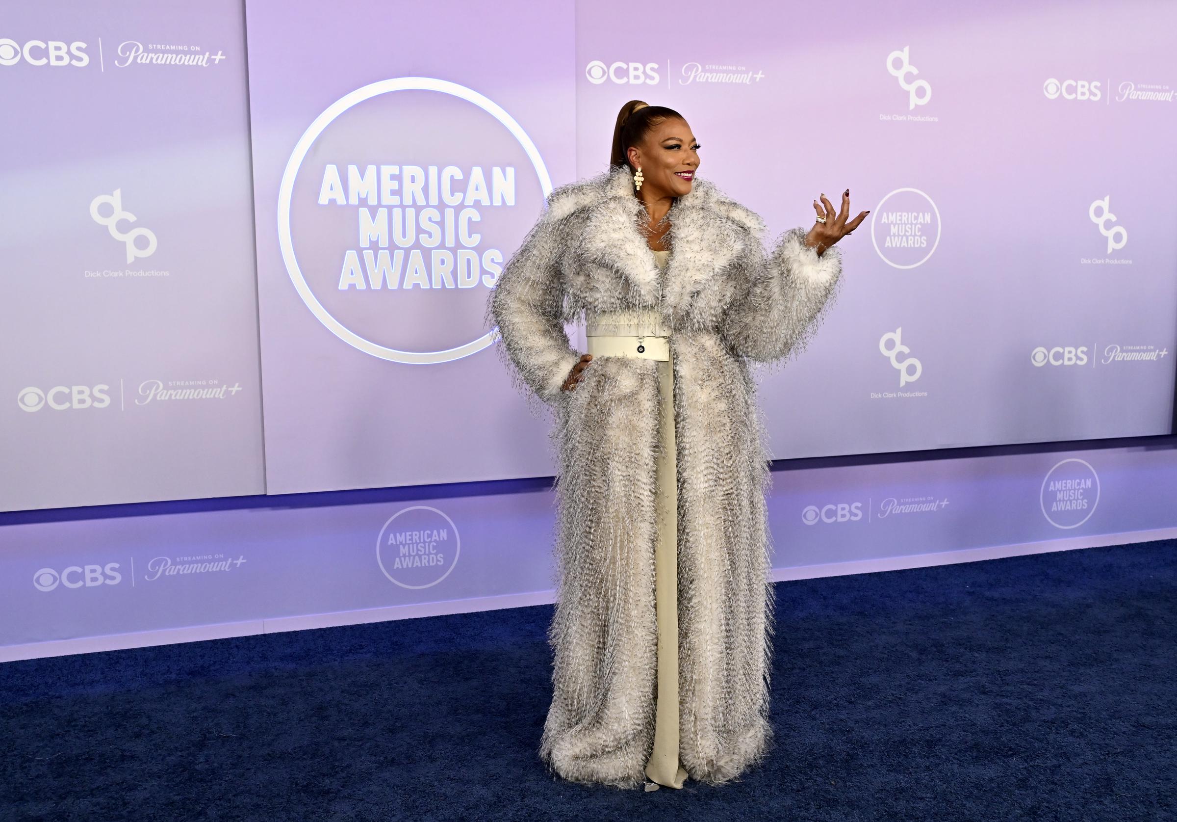 Queen Latifah strikes a confident pose on the blue carpet as CBS and American Music Awards logos shine across the lavender backdrop behind her.